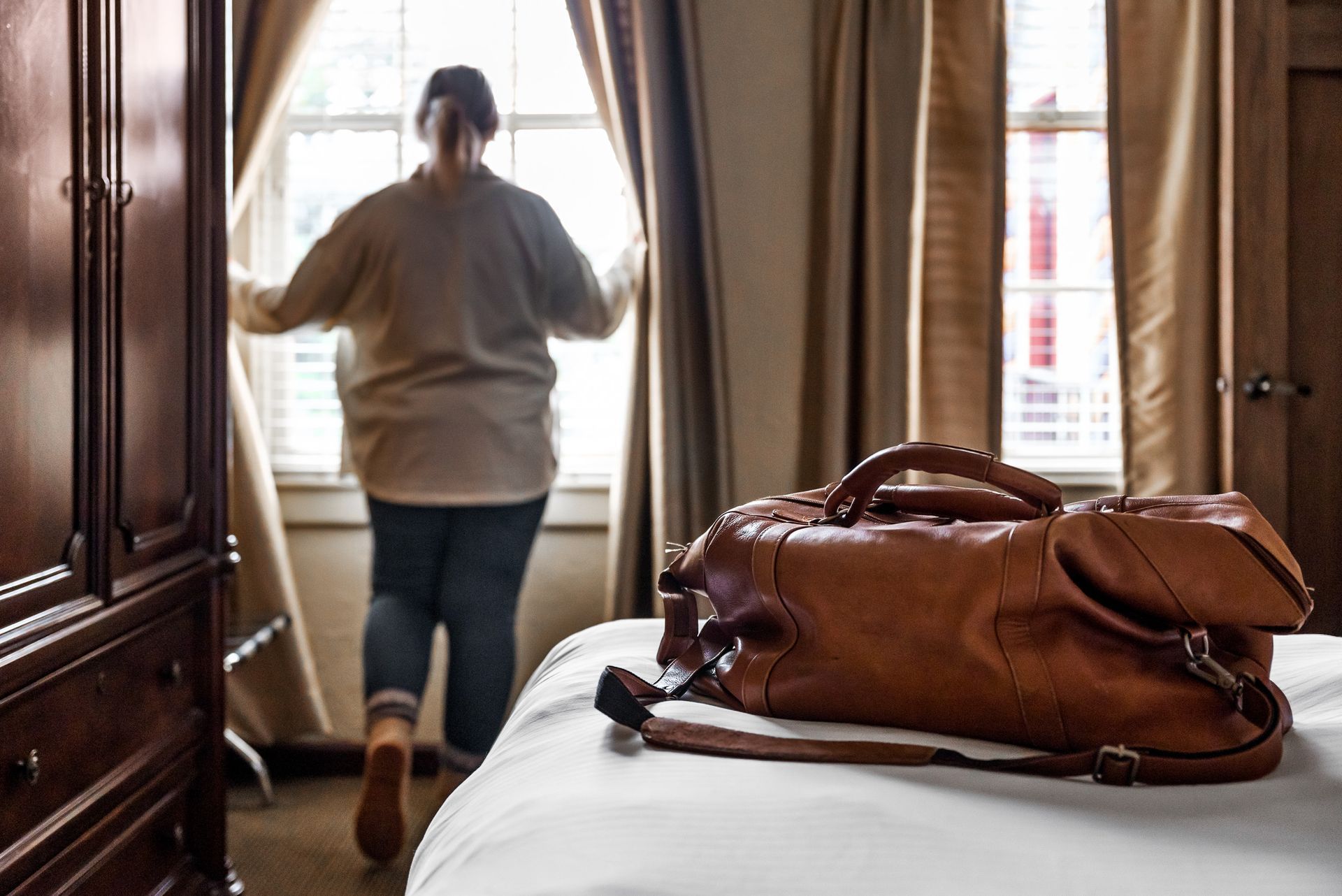 Woman looking out a window in a hotel room with a leather bag on the bed.