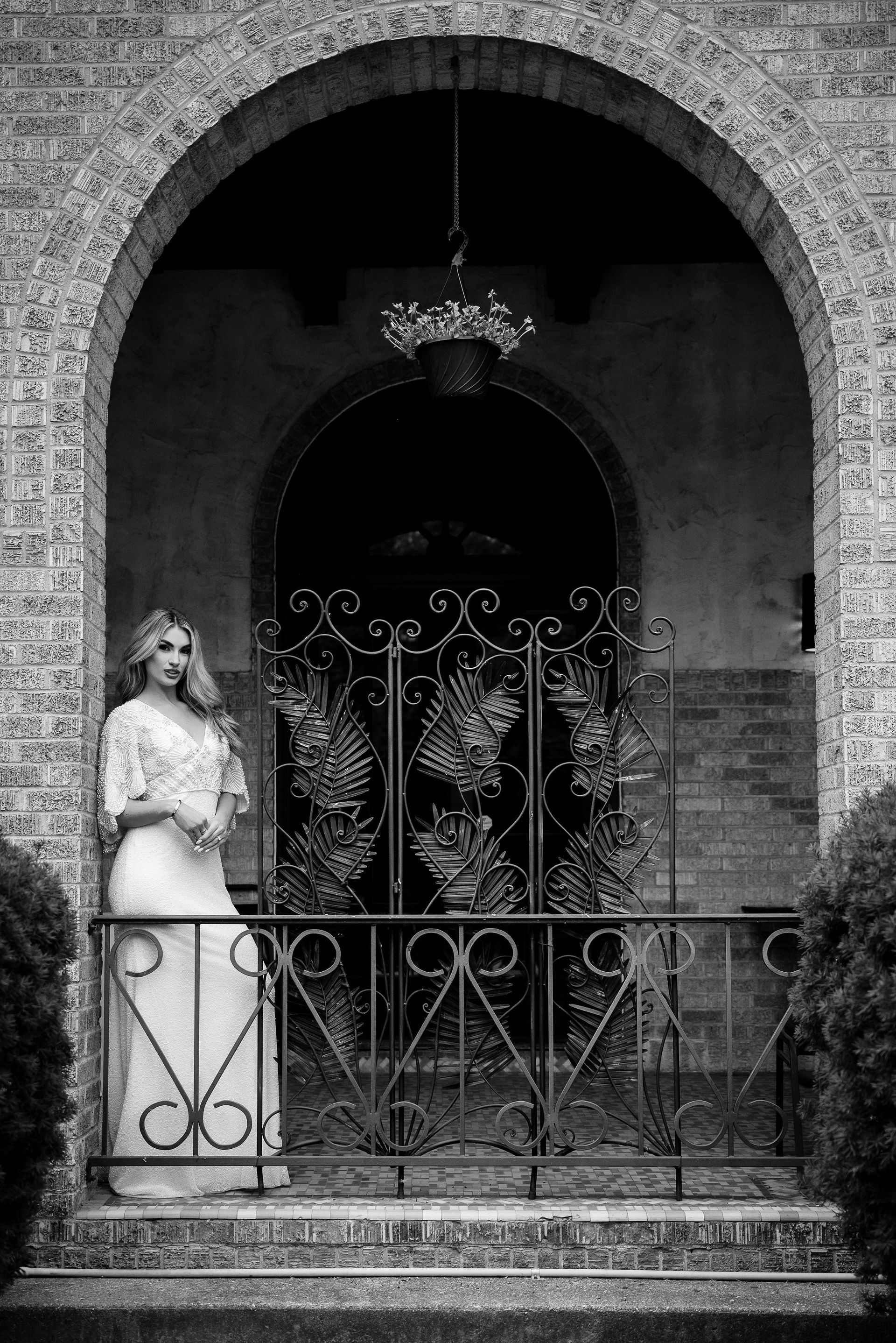 Bride in wedding dress stands inside arched doorway with ornate gate. Black and white.