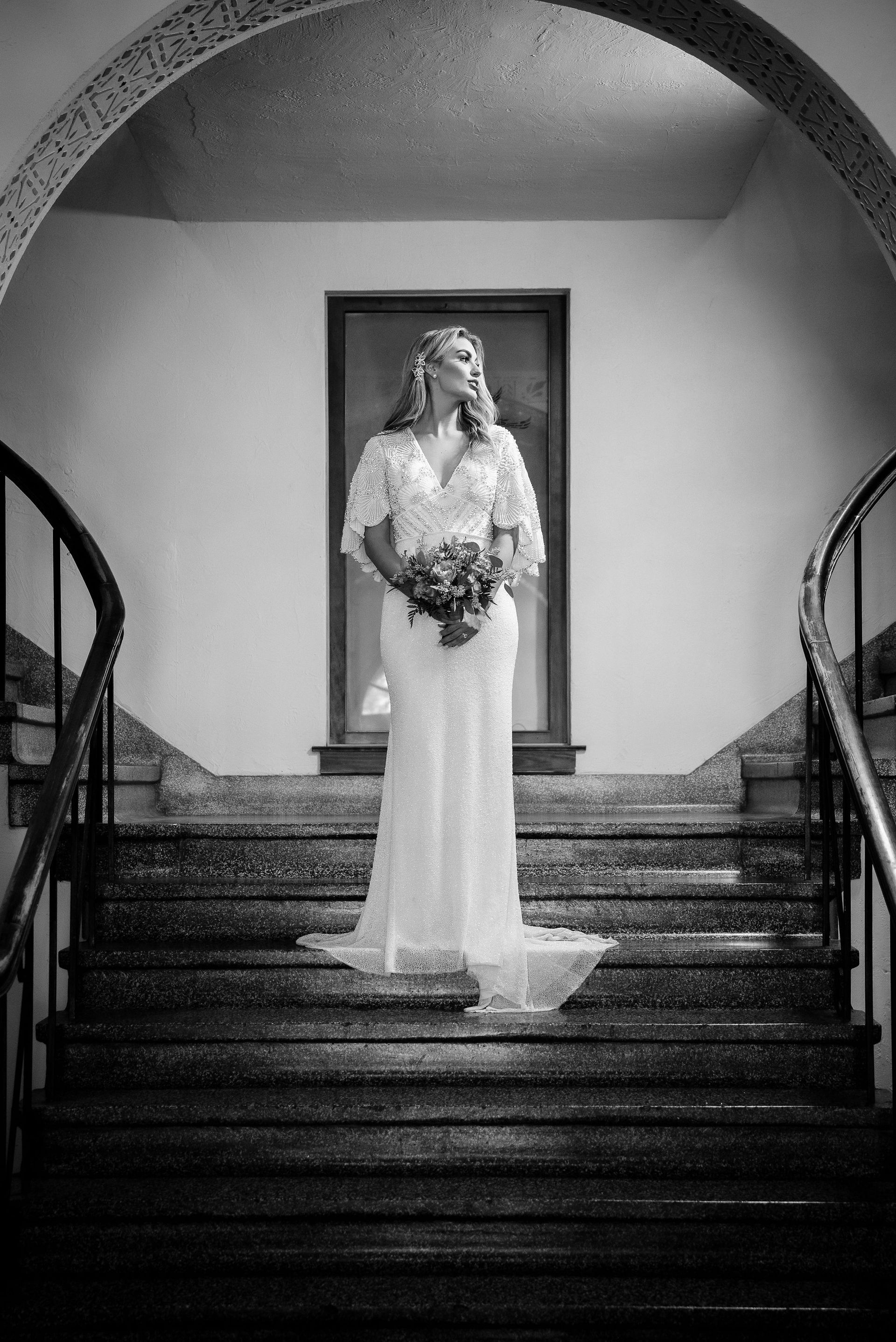 Bride in a white dress stands on stairs, holding flowers.