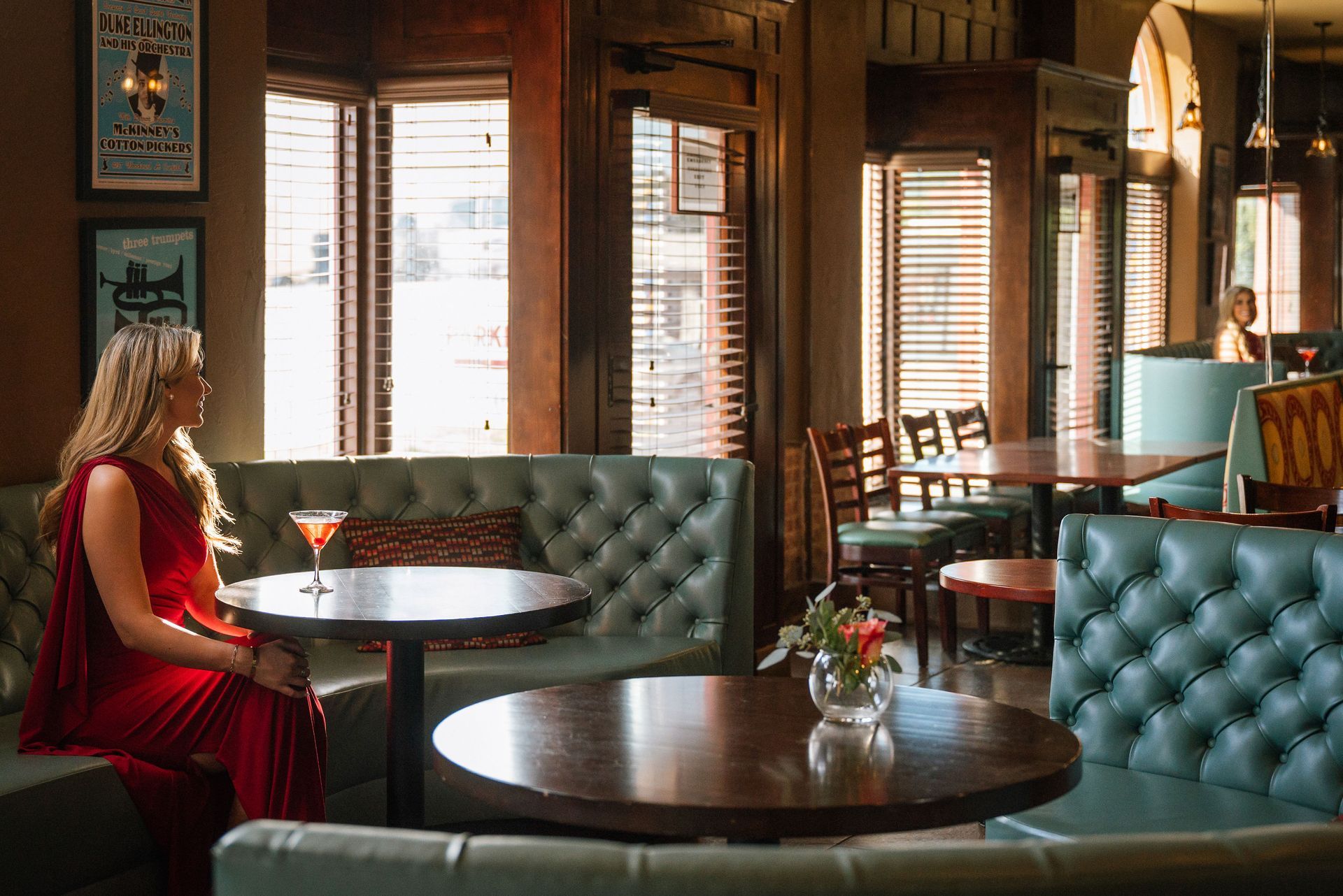 Woman in red dress sitting at a table in a dimly lit bar with green booths, waiting.