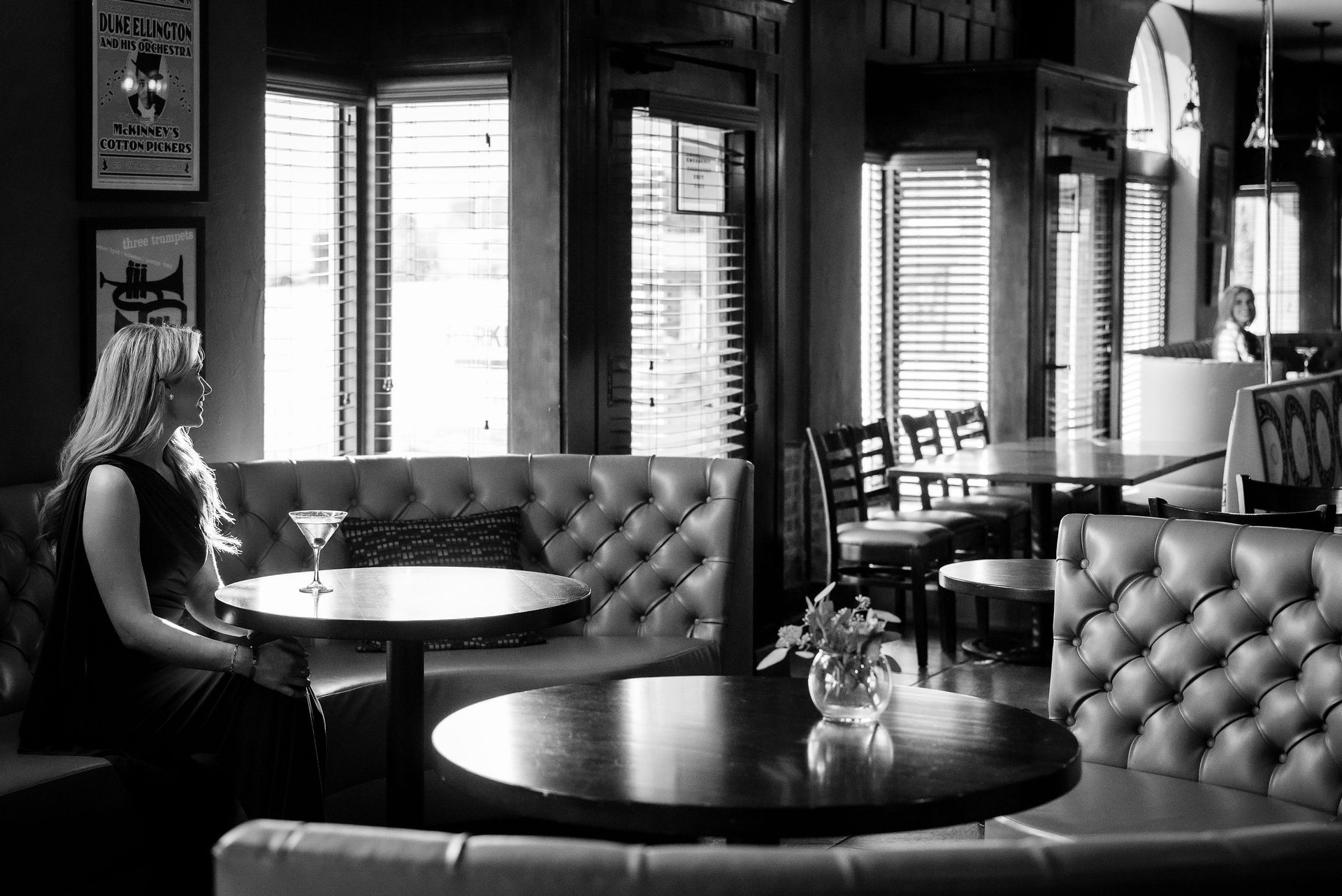 Woman sits alone in a dimly lit bar with a martini. Leather booths and round tables, open shutters.
