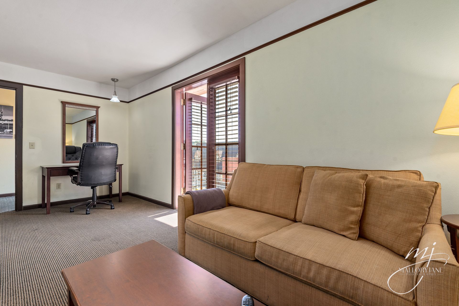 A hotel room with a sofa, desk, and balcony. Beige walls, brown furniture, patterned carpet.