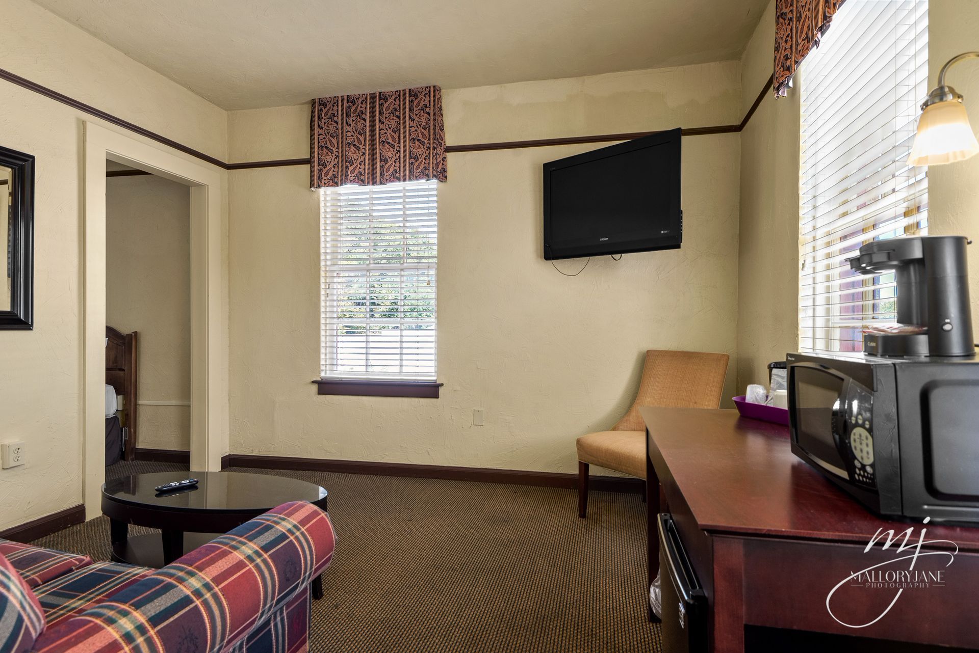 Hotel room with plaid couch, TV, windows with blinds, desk with microwave and coffee maker.