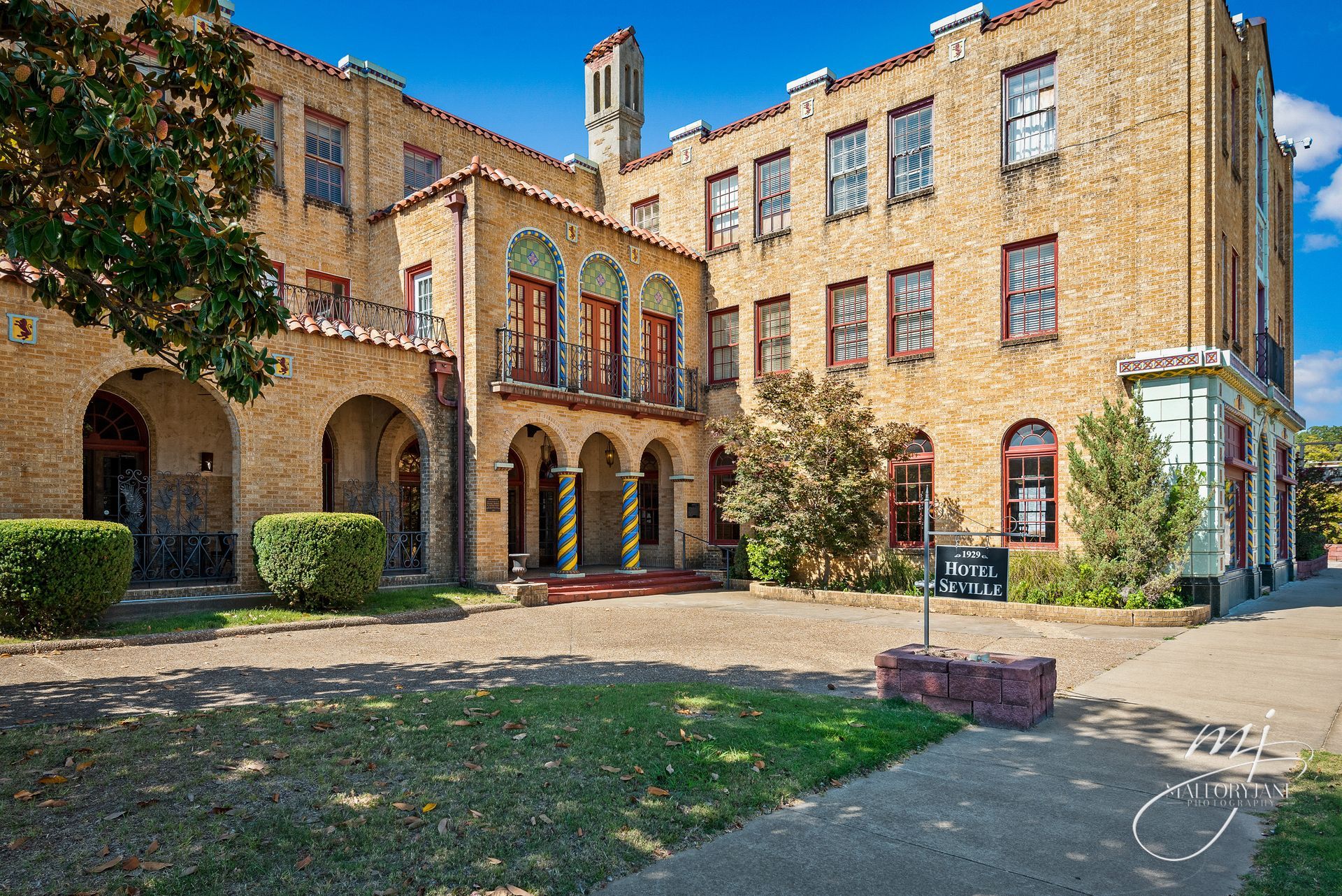 Beige brick building with arched walkway and courtyard; green bushes, sky, and sign visible.