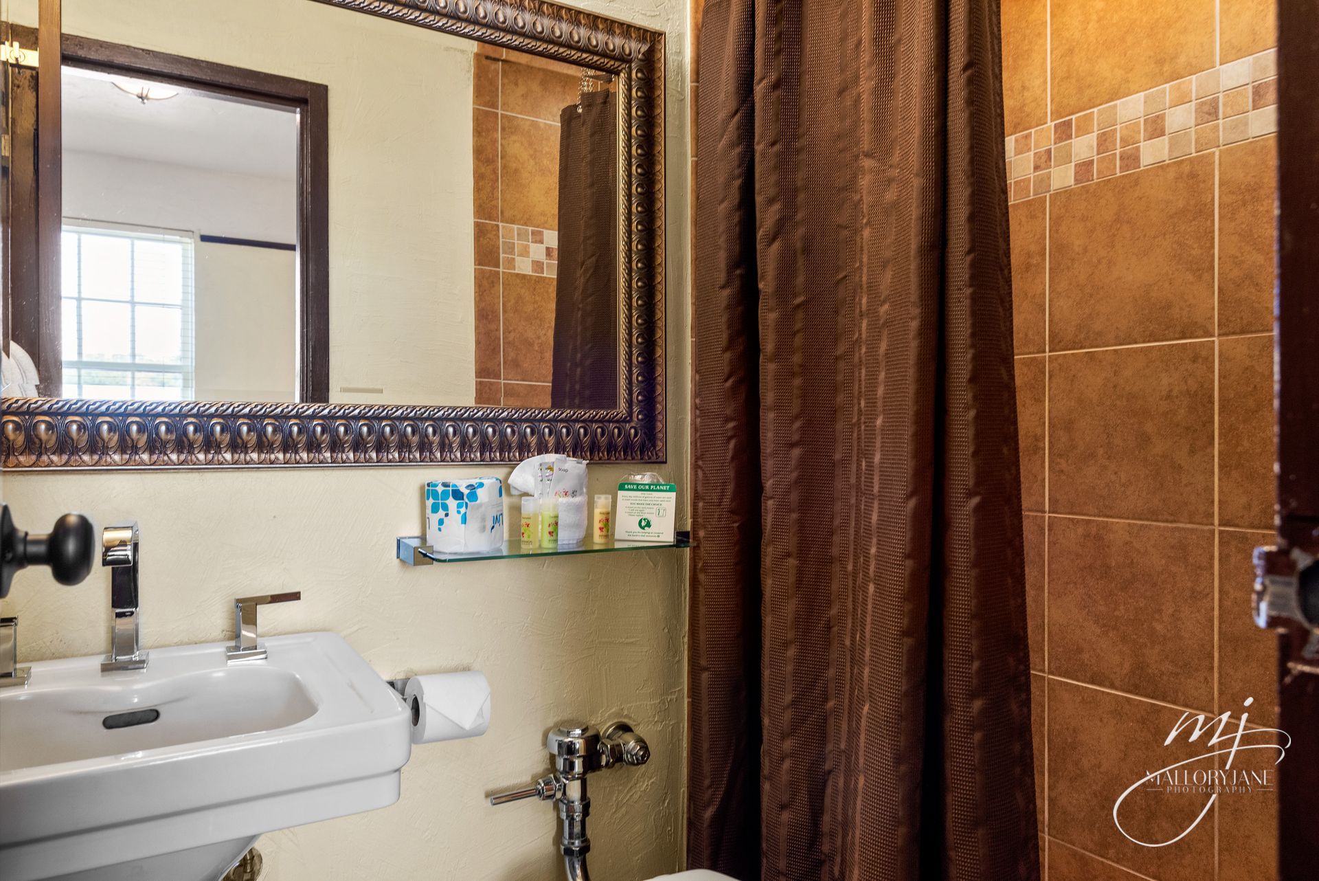 Bathroom with brown tiled walls, dark shower curtain, ornate mirror, and white sink.
