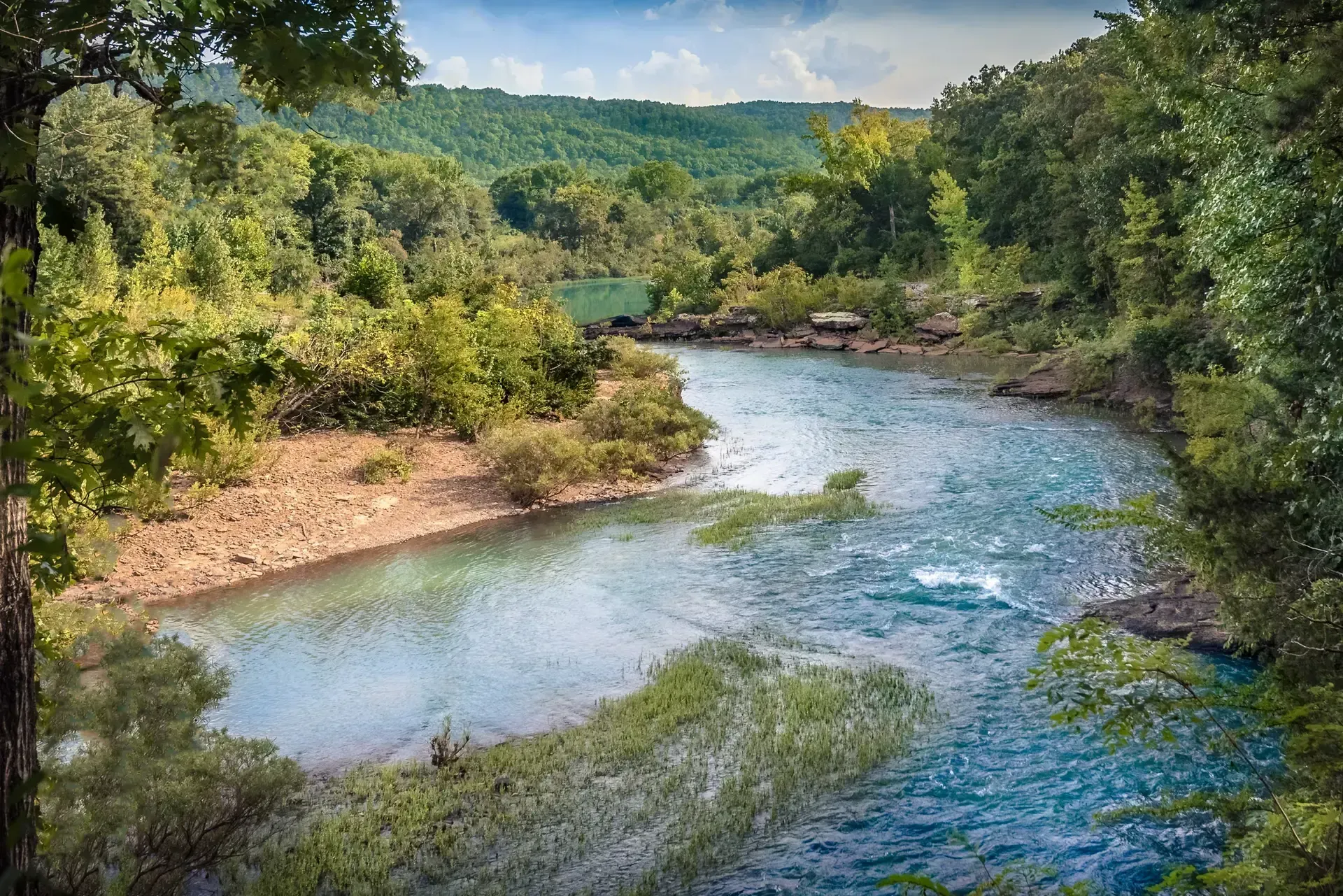 A winding river flows through a lush green forest with a glimpse of a small structure along the riverbank.