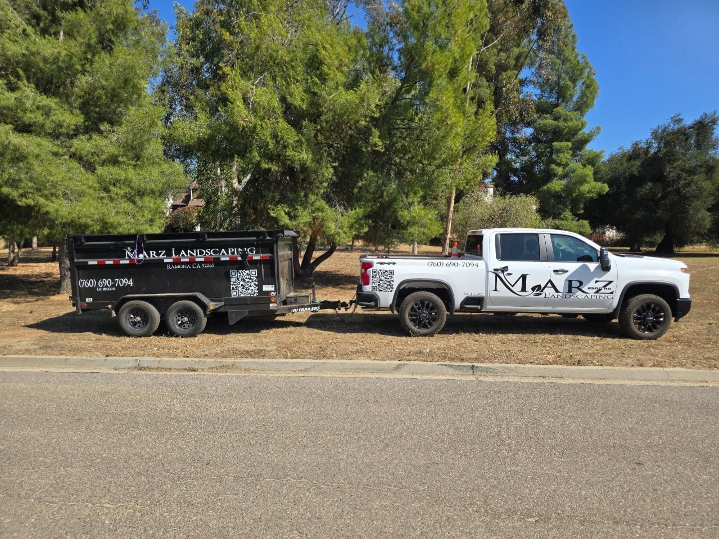 White truck pulling a black dump trailer on a road next to trees; company logo visible.