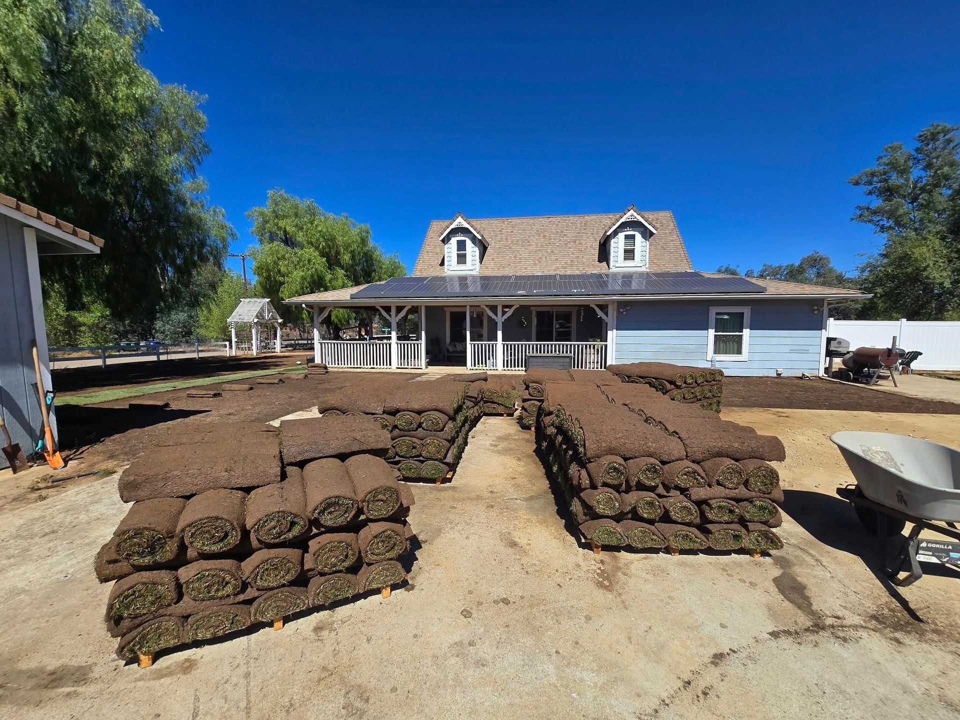 Sod rolls stacked in front of a blue house with solar panels on the roof under a bright blue sky.