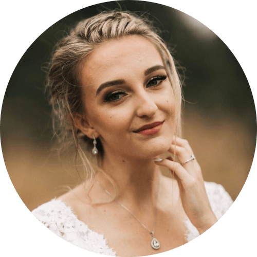 A woman in a white dress and necklace is smiling and touching her face.