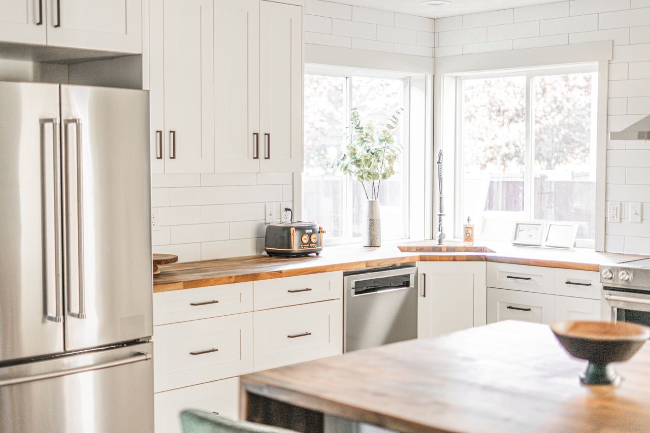 A kitchen with stainless steel appliances , white cabinets , wooden counter tops , and a large window.