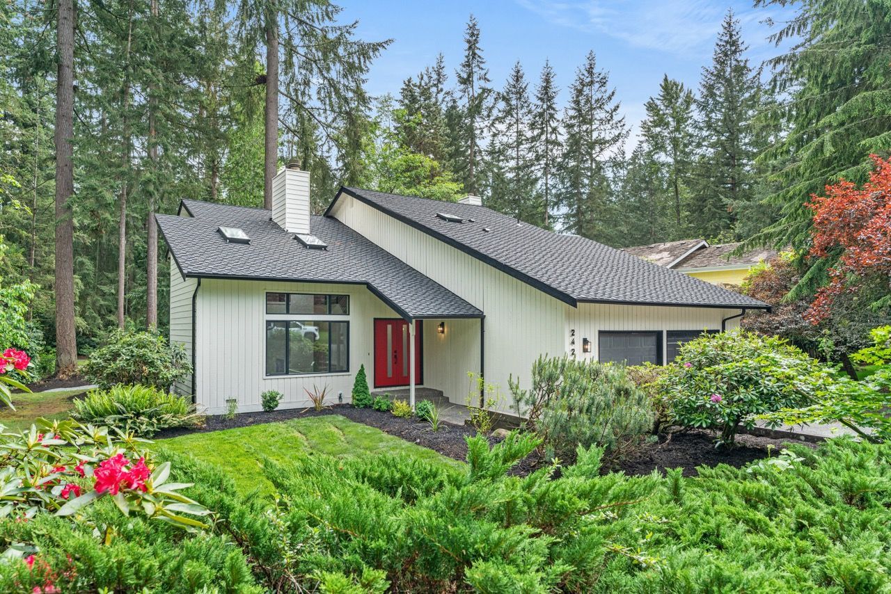 A white house with a red door is surrounded by trees and bushes.