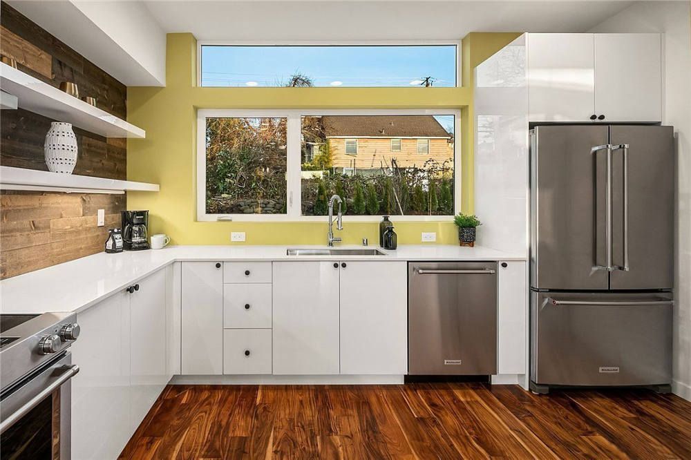 A kitchen with white cabinets , stainless steel appliances , hardwood floors and a large window.