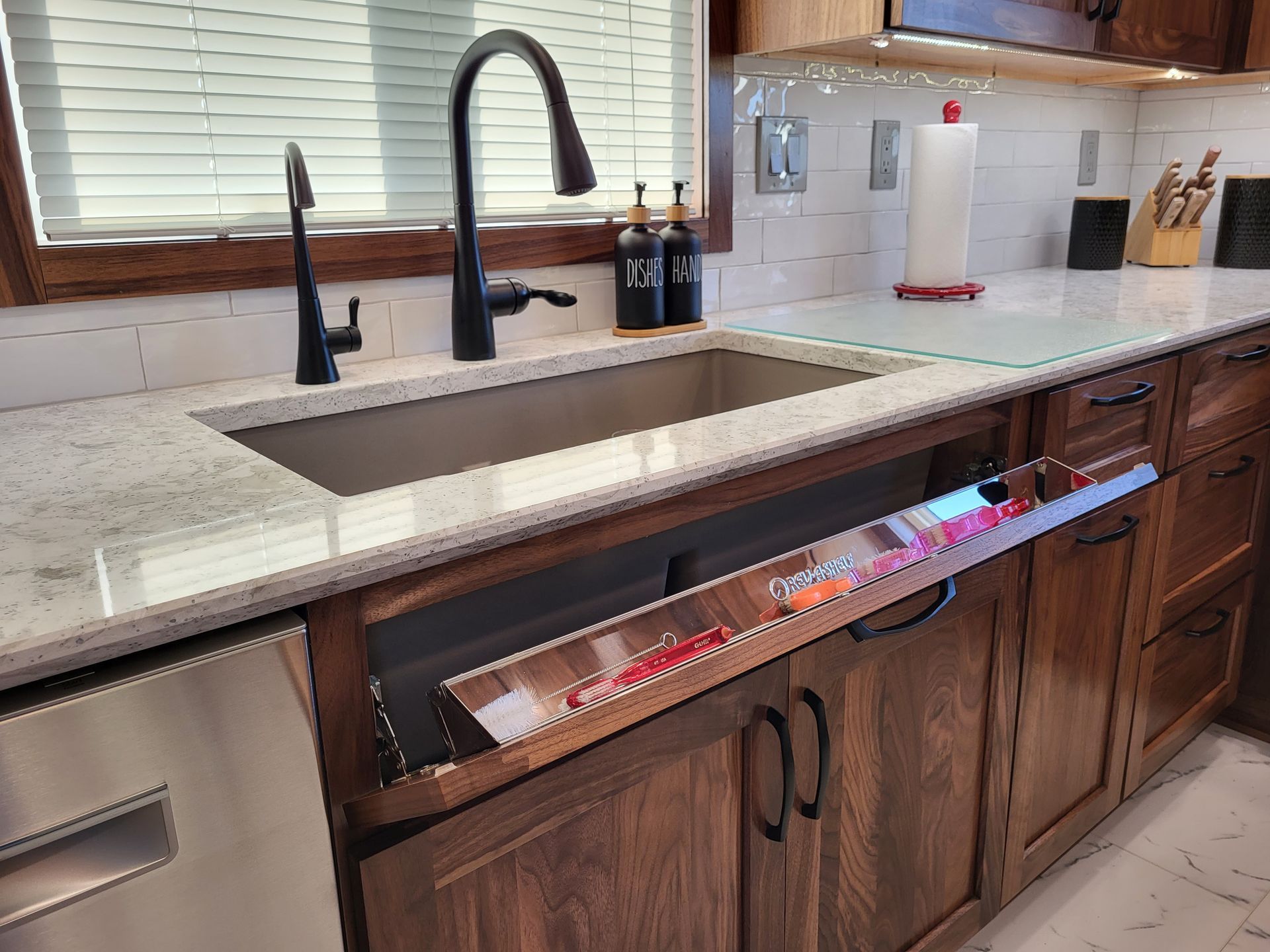 Kitchen sink with a pull-out knife drawer. Stainless steel sink, wooden cabinets, and black faucet.