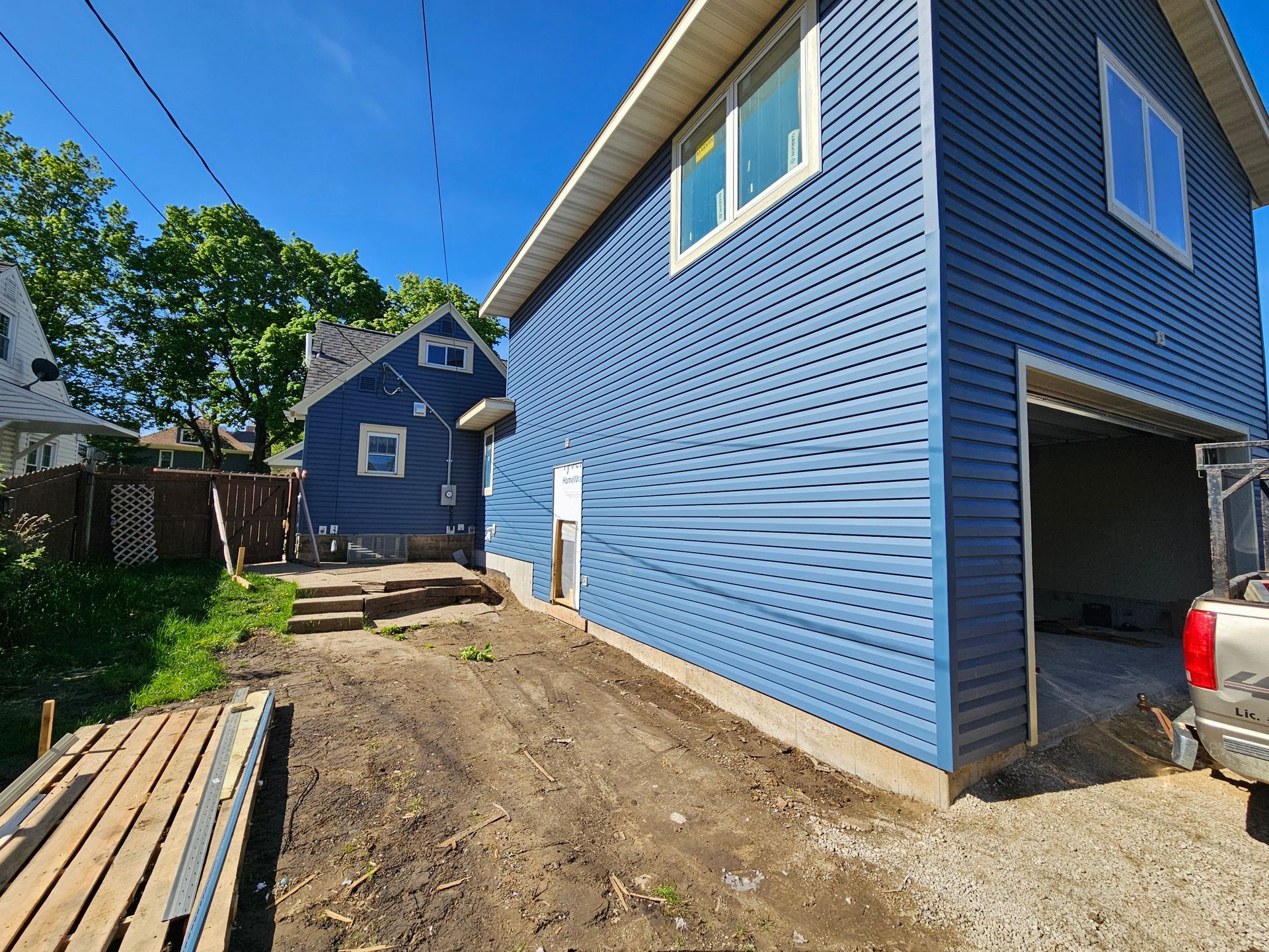 Blue house with open garage, dirt driveway, and steps leading to another blue house on a sunny day.