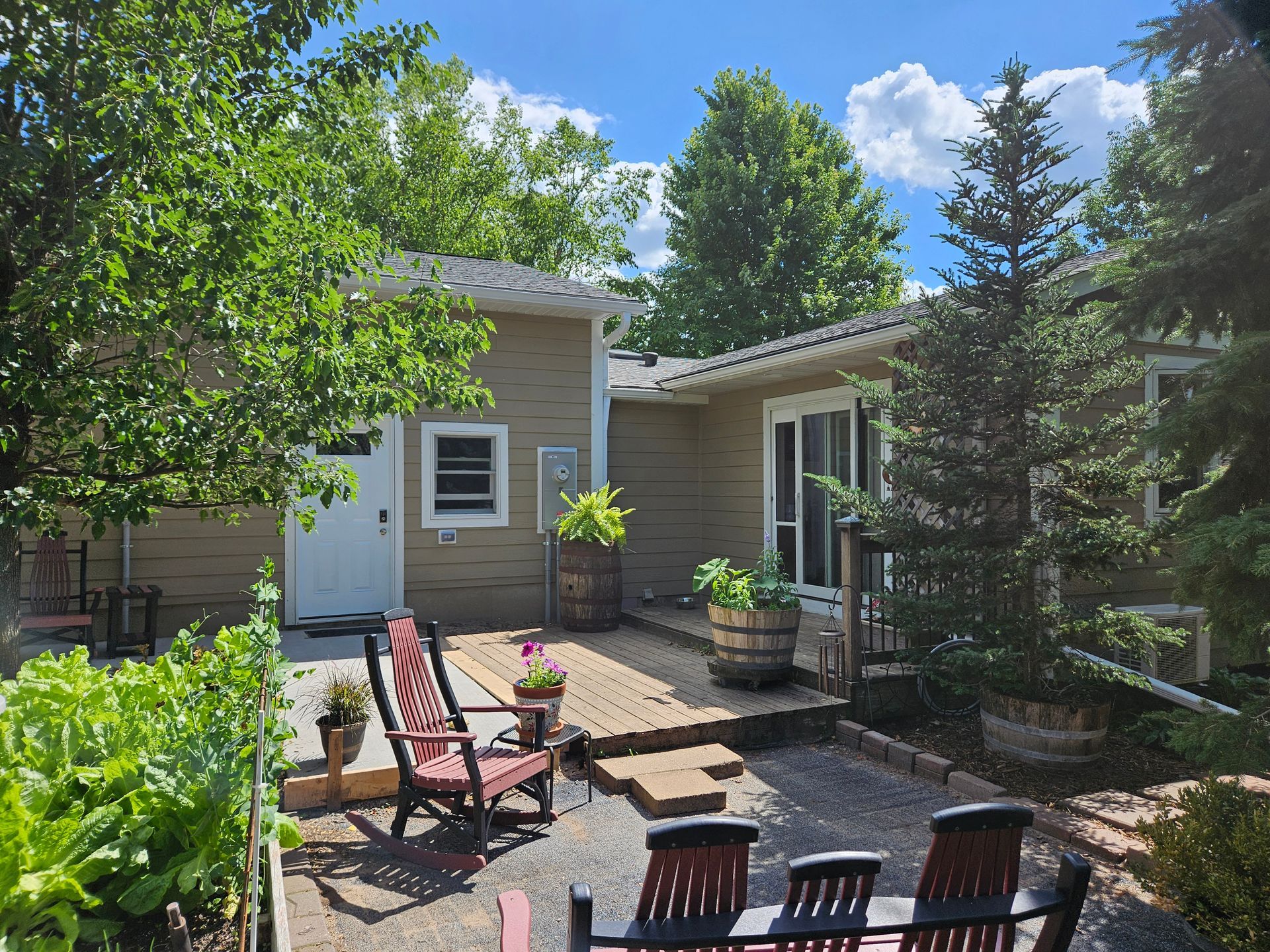 Backyard deck with chairs, potted plants, and a small building with a white door, sunny day.