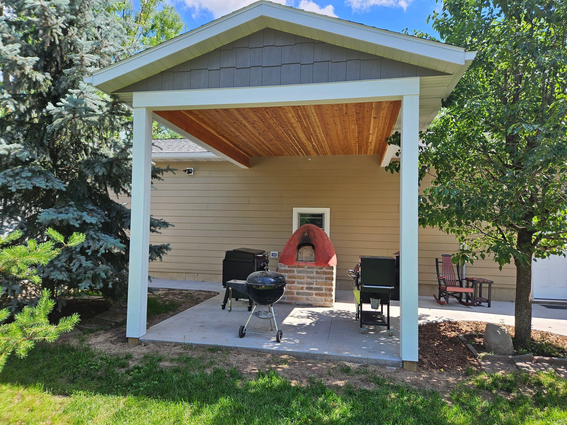 Covered patio with grill, red brick oven, and rocking chairs next to a beige building.