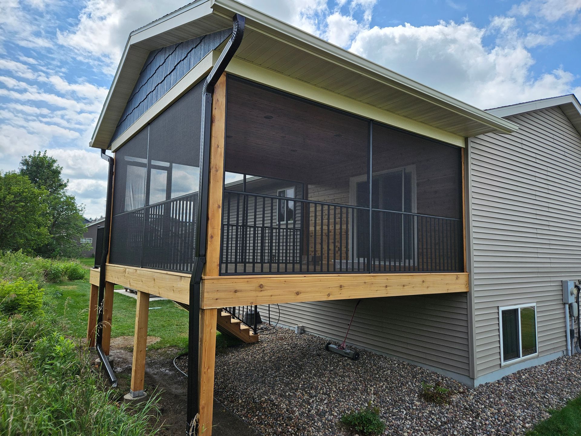 Screened-in porch with wooden deck, black screen, attached to a house with siding. Blue sky visible.