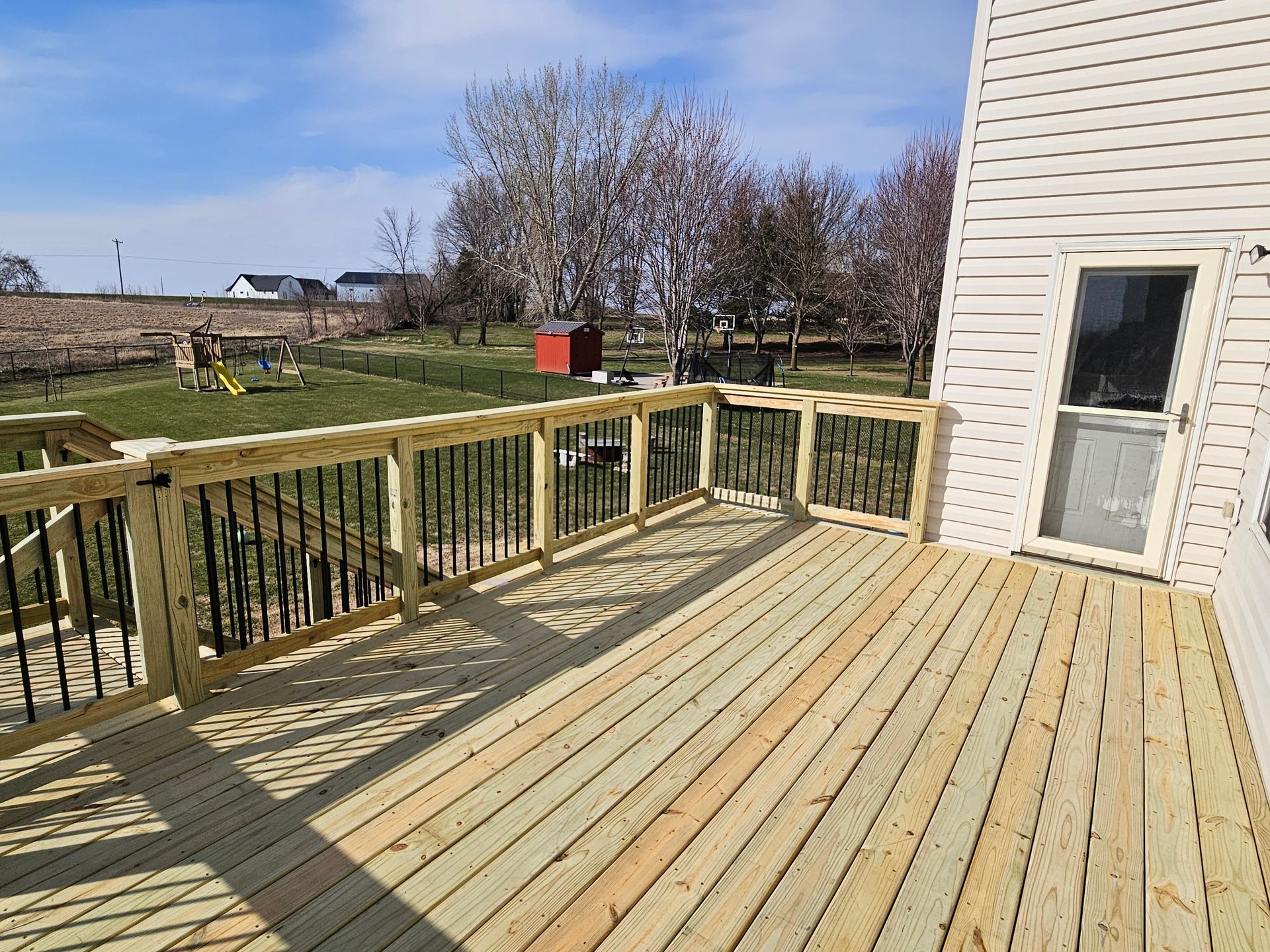 Wooden deck with black railing next to a house, overlooking a yard with a playground.