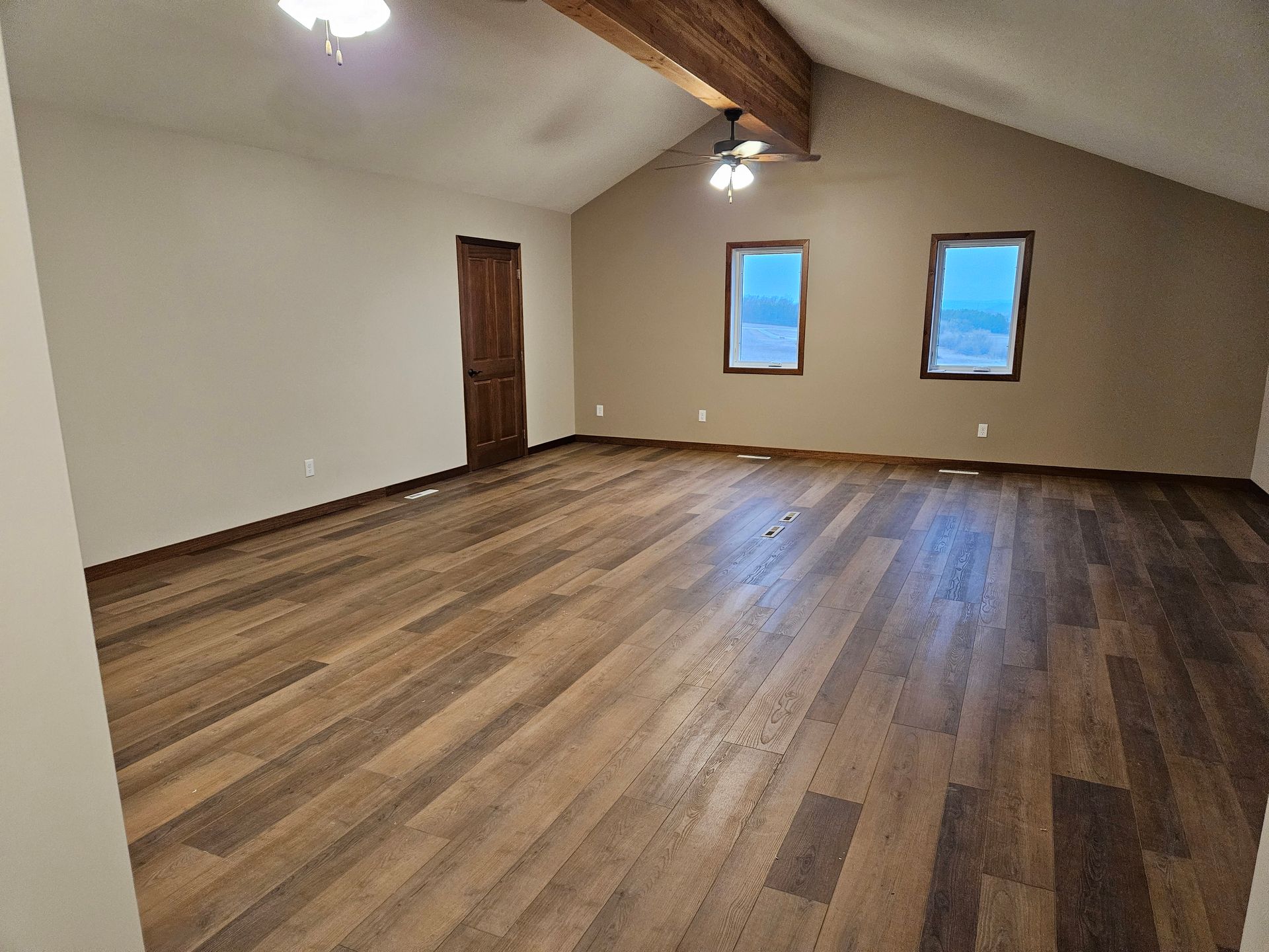 Empty living room with wood floor, vaulted ceiling, wood beam, windows, and brown walls.
