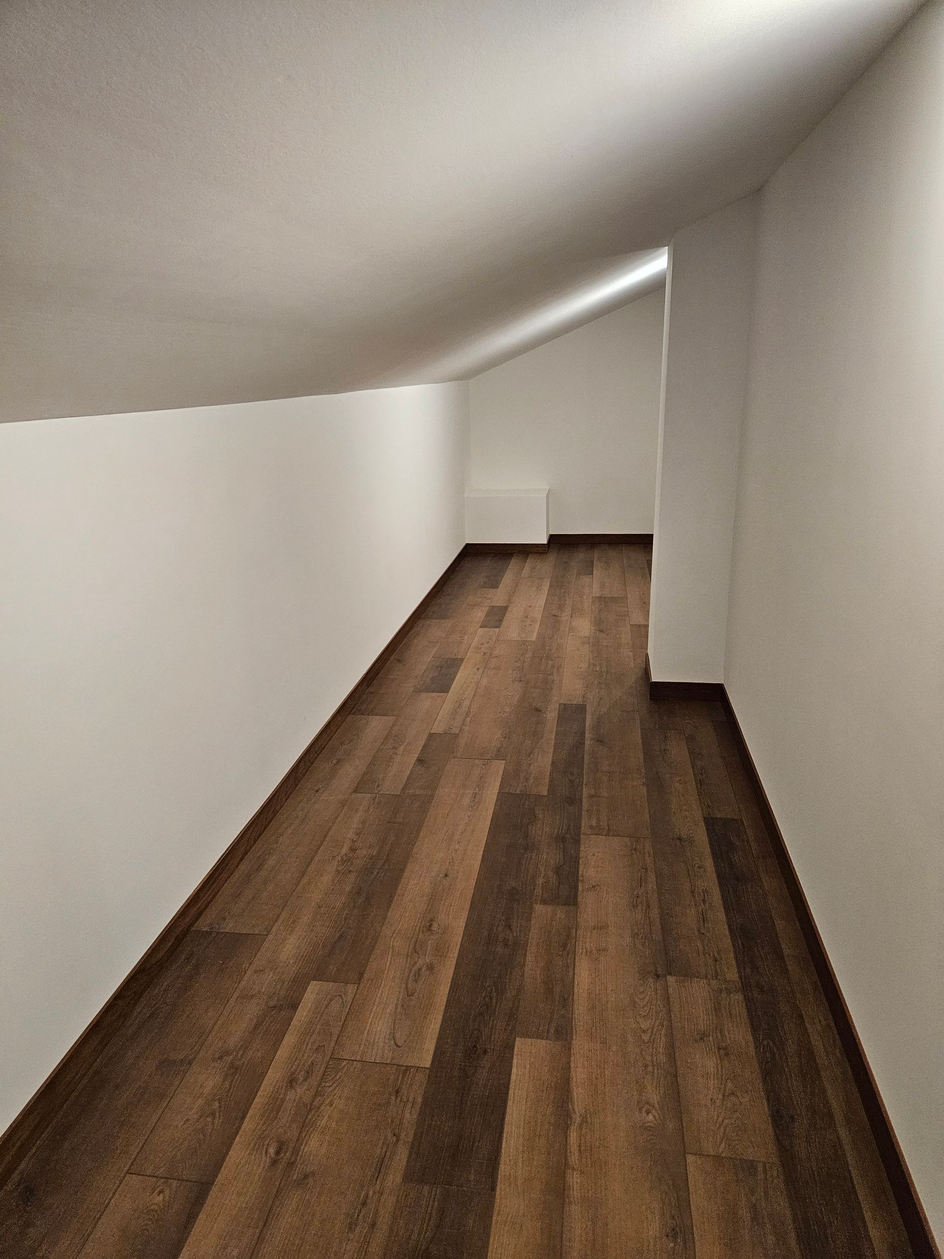 Narrow attic room with slanted ceiling, wood-look flooring, and white walls.