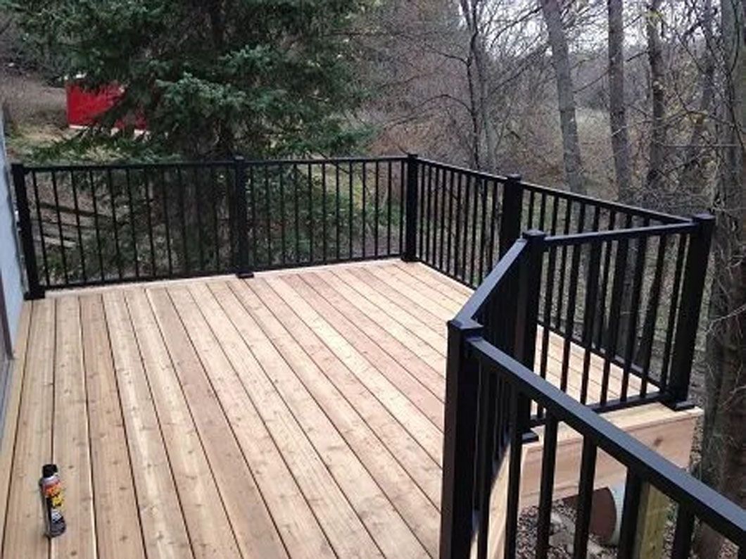 Wooden deck with black railing in a wooded setting; a red building visible in the background.