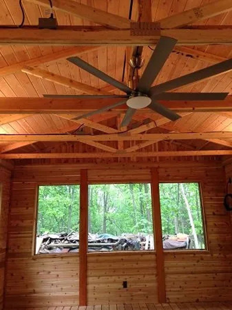 Wooden interior with large fan, windows overlooking a forest, and exposed beams.