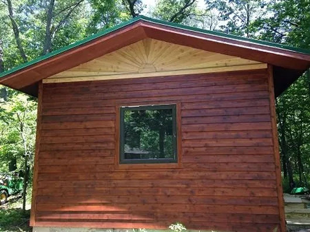 Brown cabin with a green roof and one window, surrounded by trees.