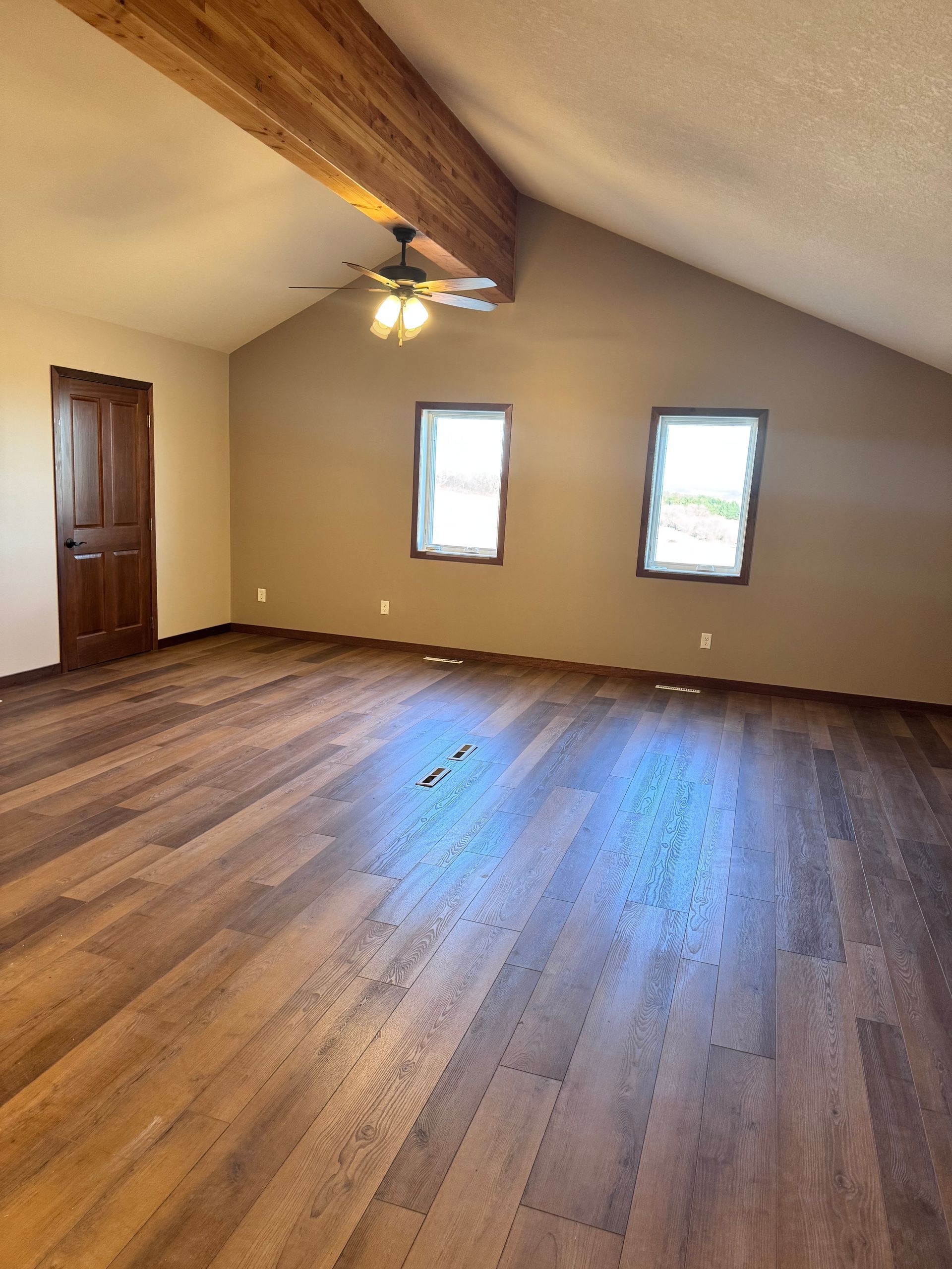 Spacious room with wood floors and brown walls, two windows, a wood door, and ceiling beam.