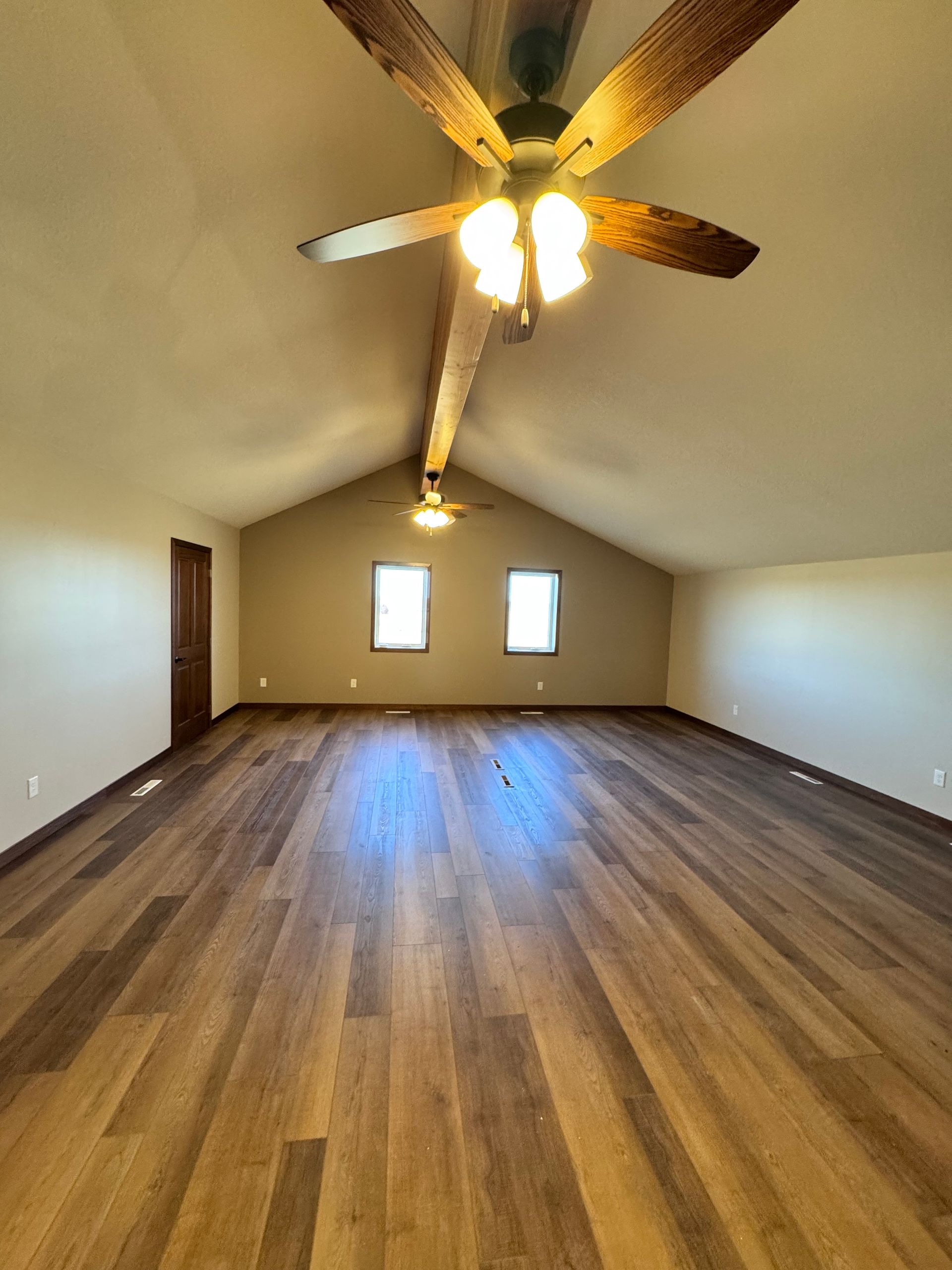 Spacious room with hardwood floors, vaulted ceiling, ceiling fan, and two windows.