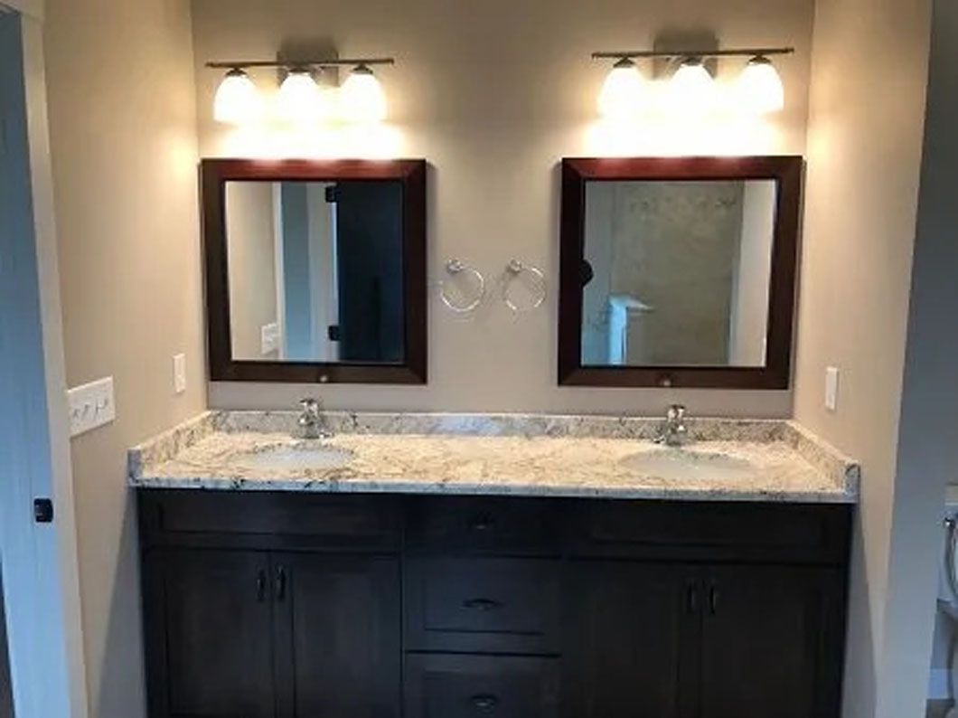 Bathroom with two sinks, mirrors, and lights above a dark cabinet against a light-colored wall.