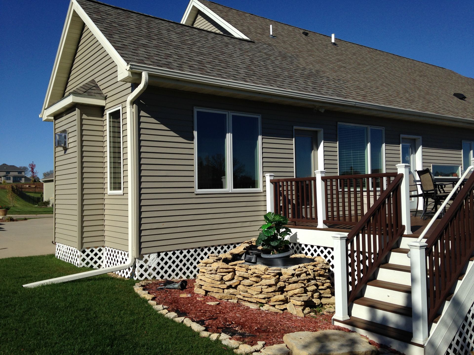 Tan house with brown deck, white lattice, and small rock fountain on green lawn.