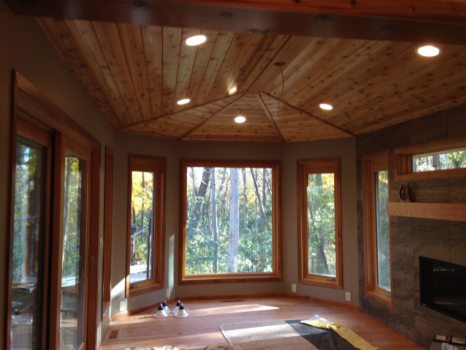 Sunroom interior with wood ceiling, windows, and a fireplace, overlooking a forest.