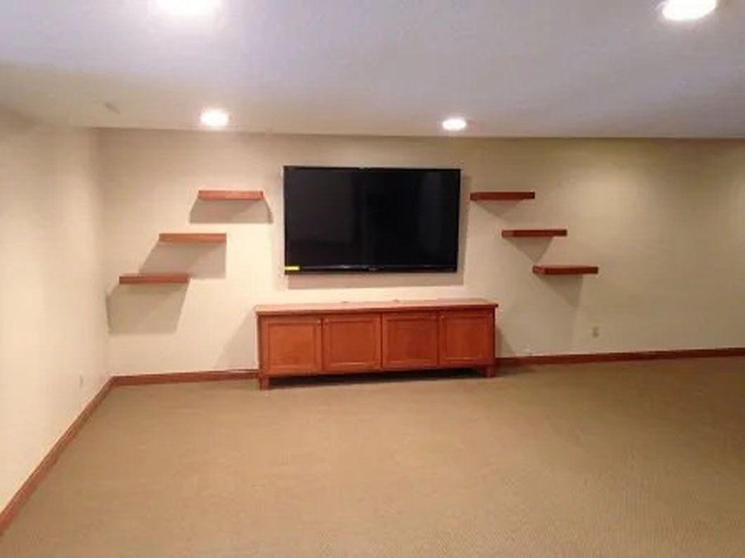 Empty room with TV mounted over a wood cabinet. Floating shelves on each side. Beige walls and carpet.