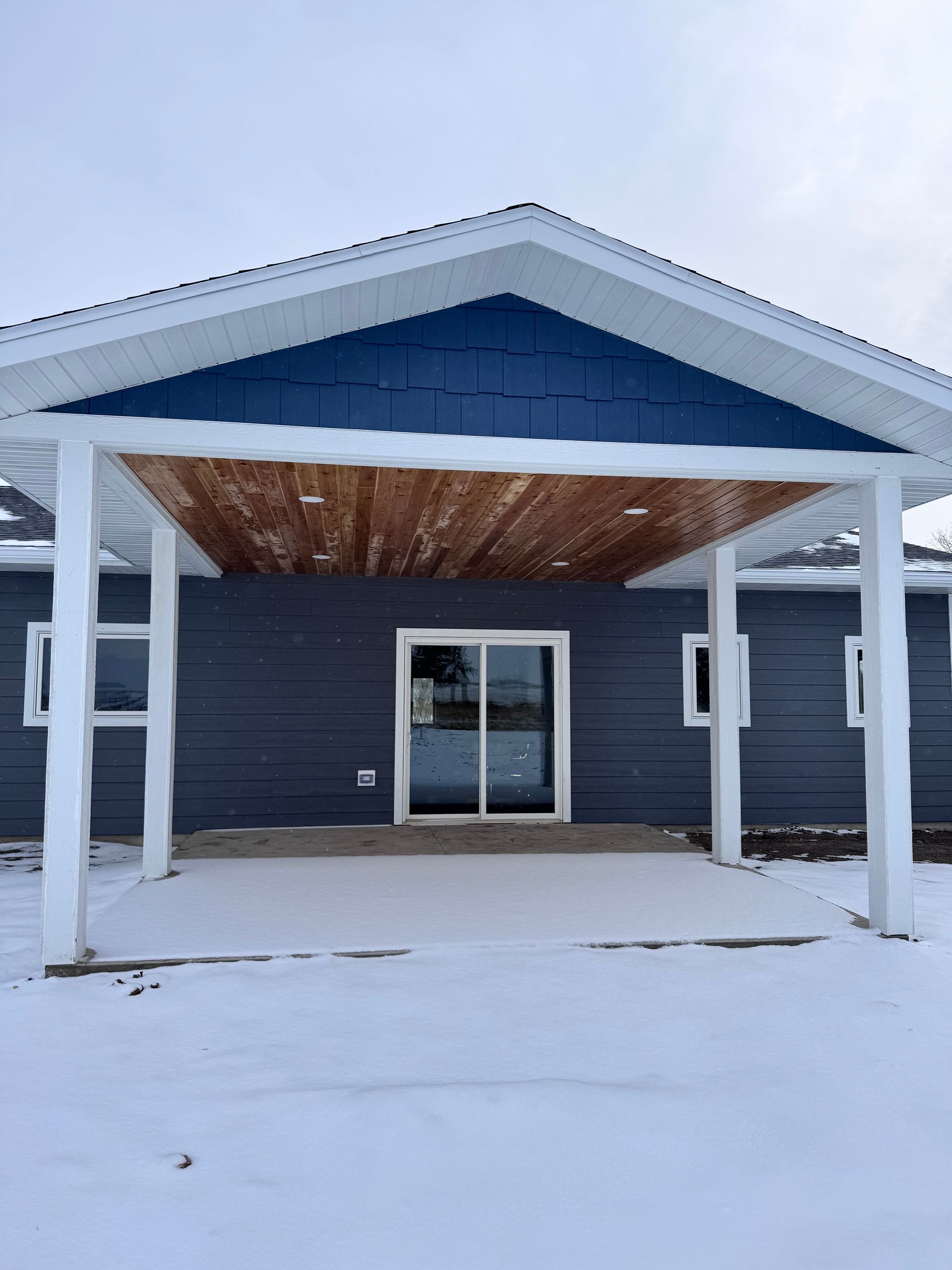 Gray building with a blue gable roof and wooden porch. Sliding glass doors and snow on the ground.