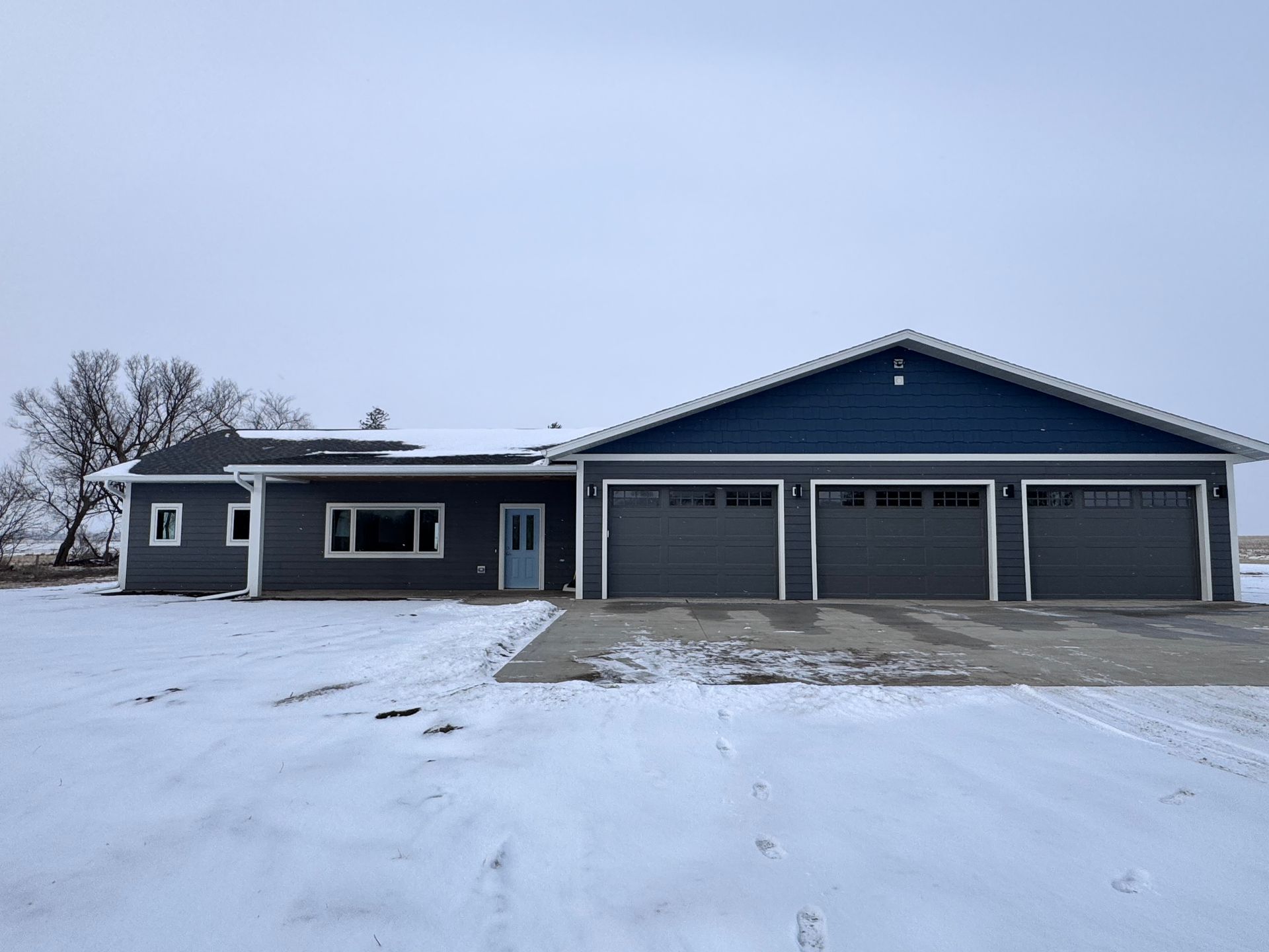 A gray and blue house with a three-car garage sits in a snowy field under a cloudy sky.