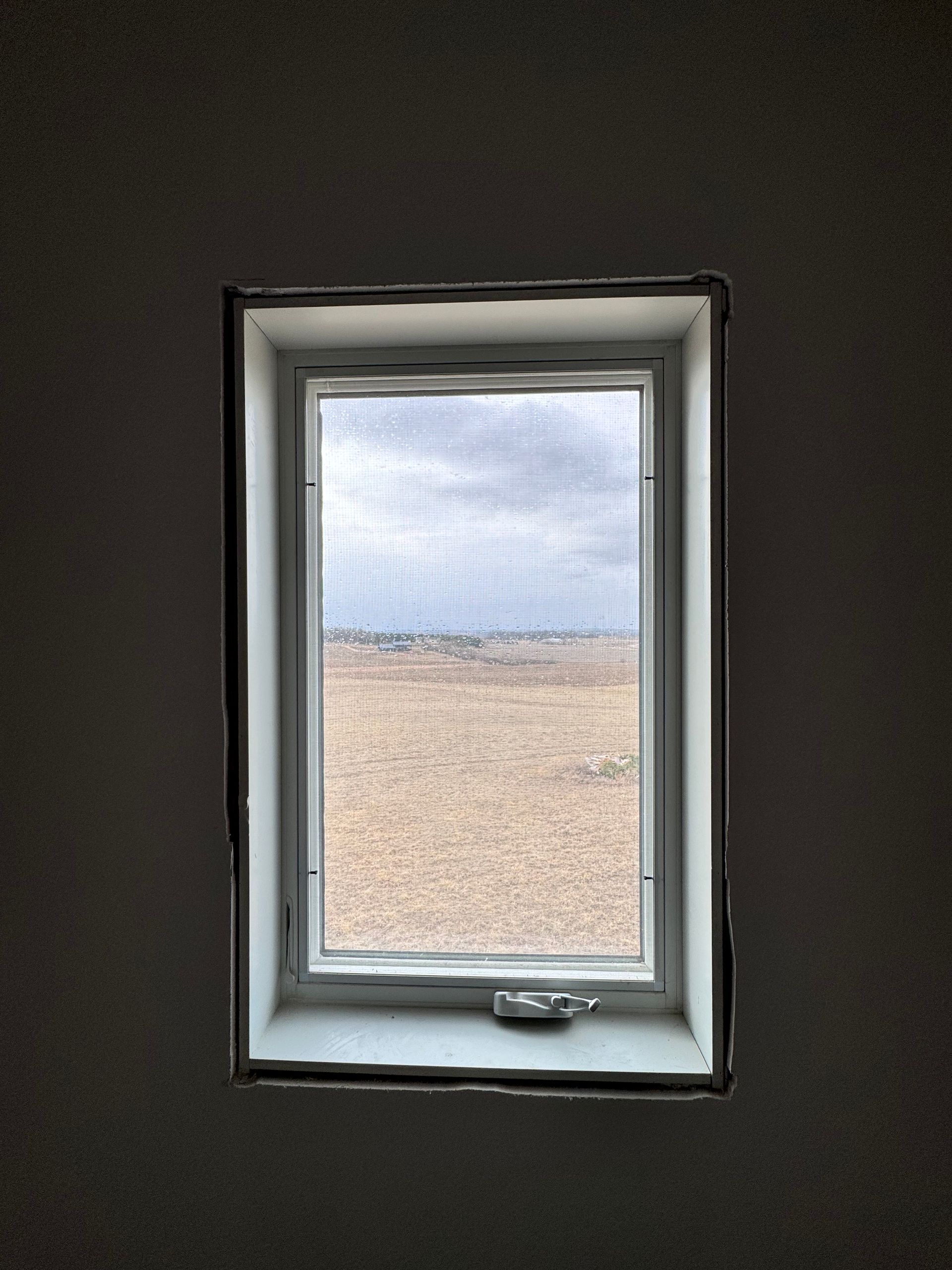Window in a dark room, looking out onto a beige field under a cloudy sky.