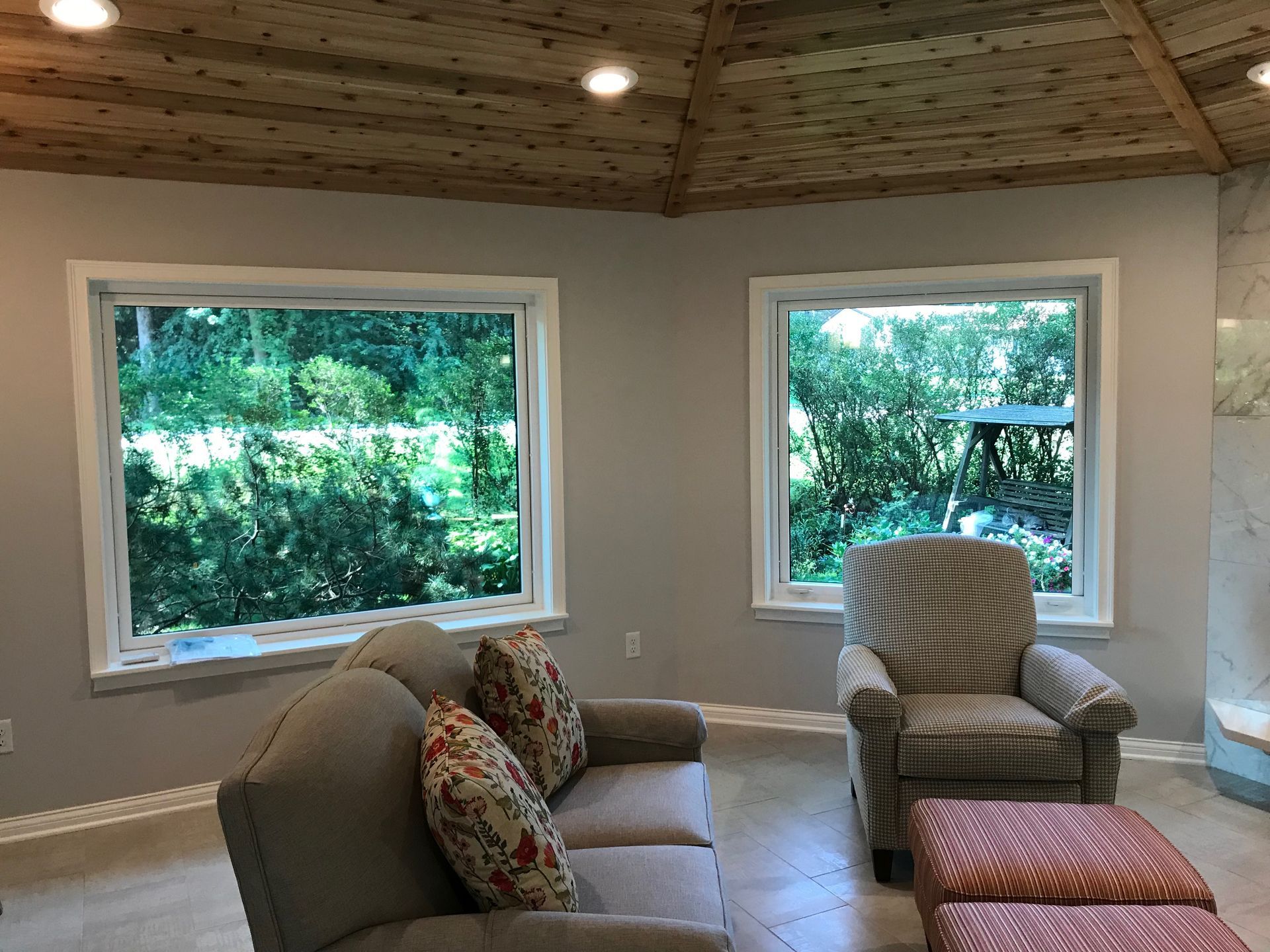 Living room with sofa and armchair facing windows overlooking greenery. Wooden ceiling, gray walls.