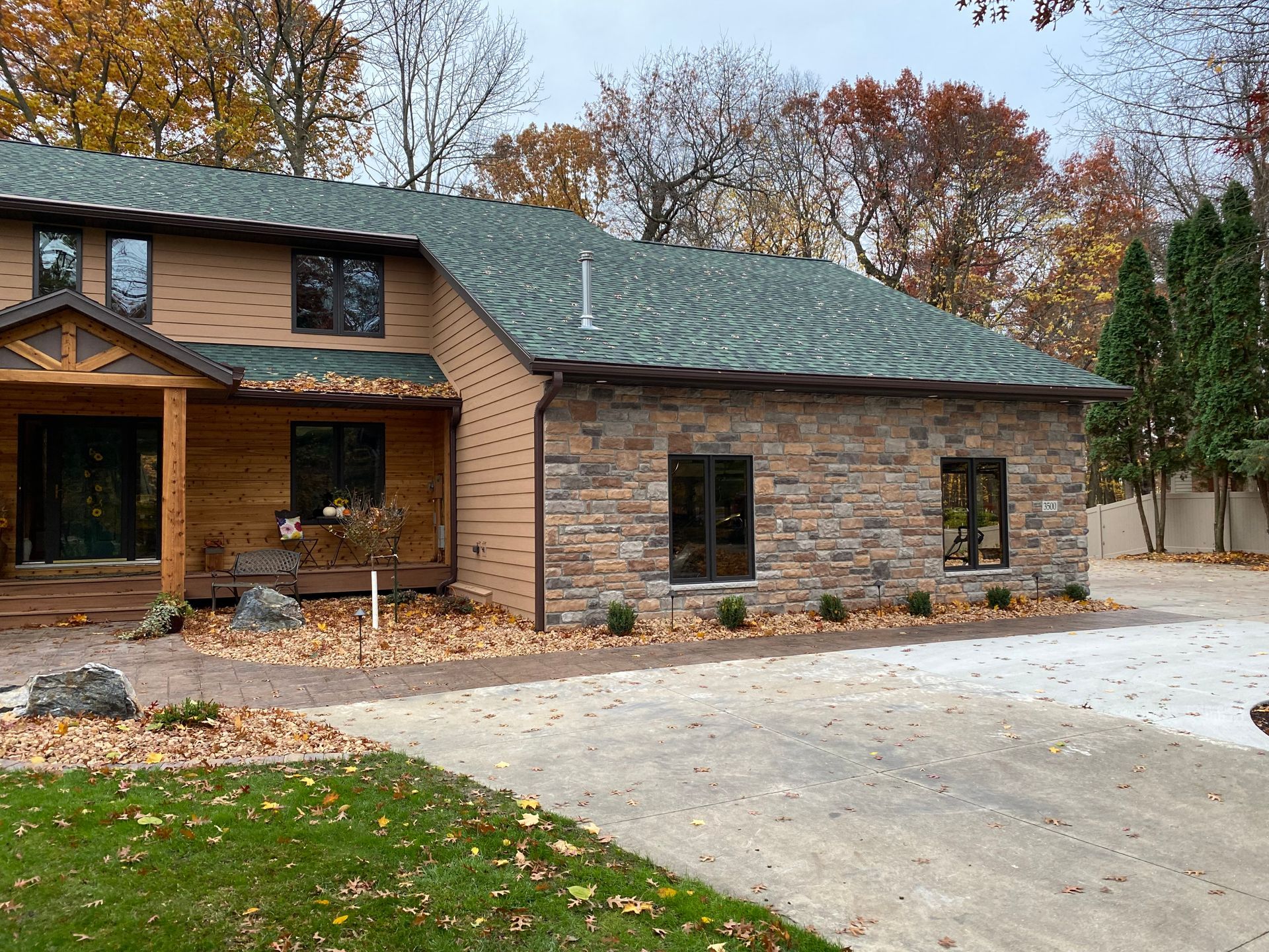 Two-story house with stone and brown siding; green roof, large windows; driveway and fall foliage.