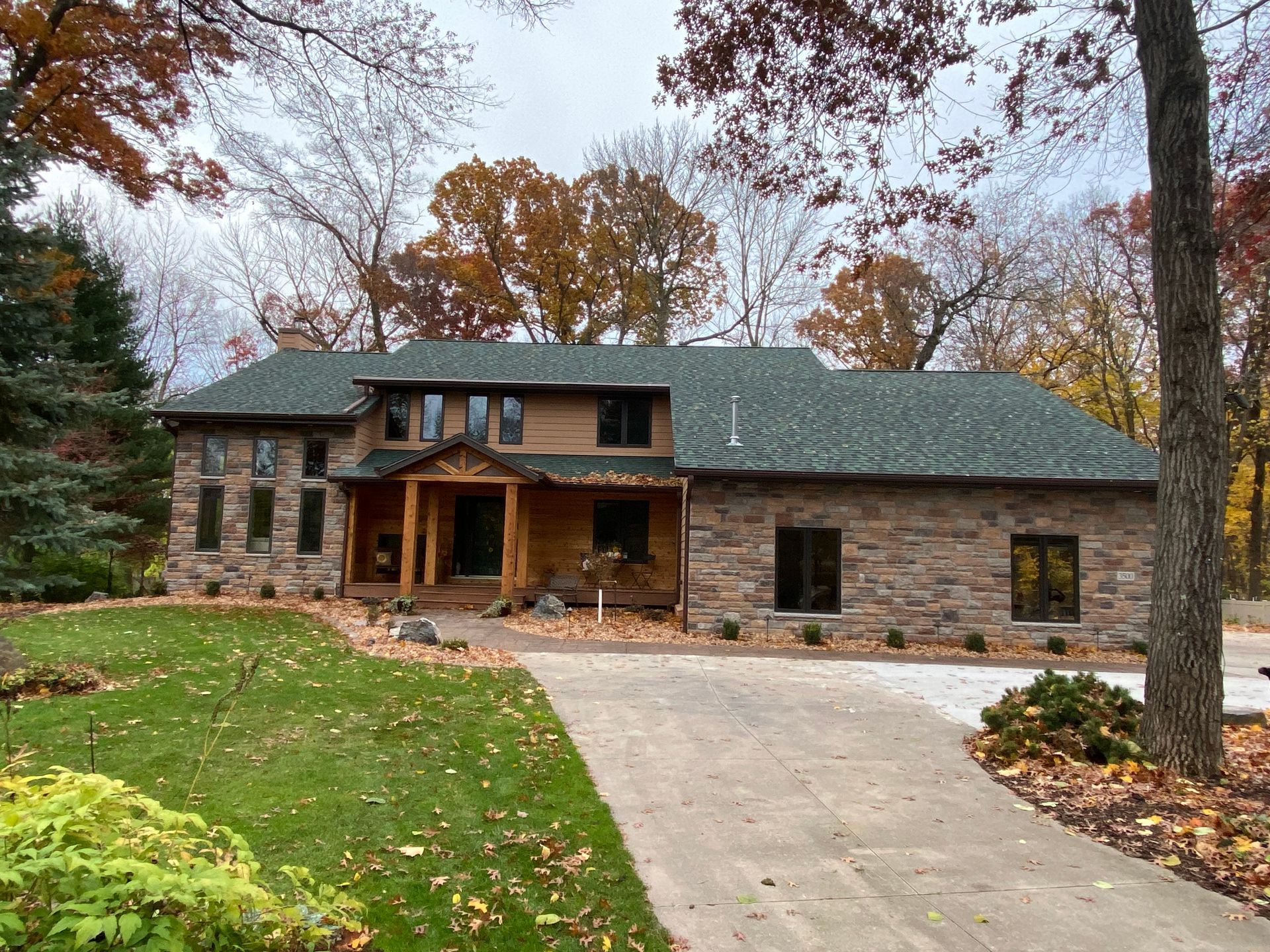 Stone house with green roof, brown wood trim, and a concrete driveway, surrounded by fall trees.