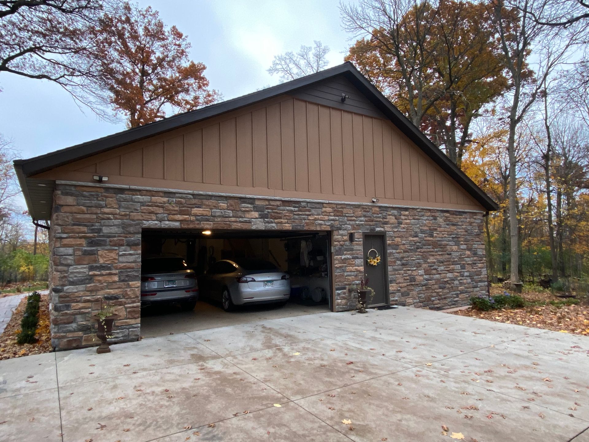 Stone garage with two cars inside, brown door, and autumn trees in the background.