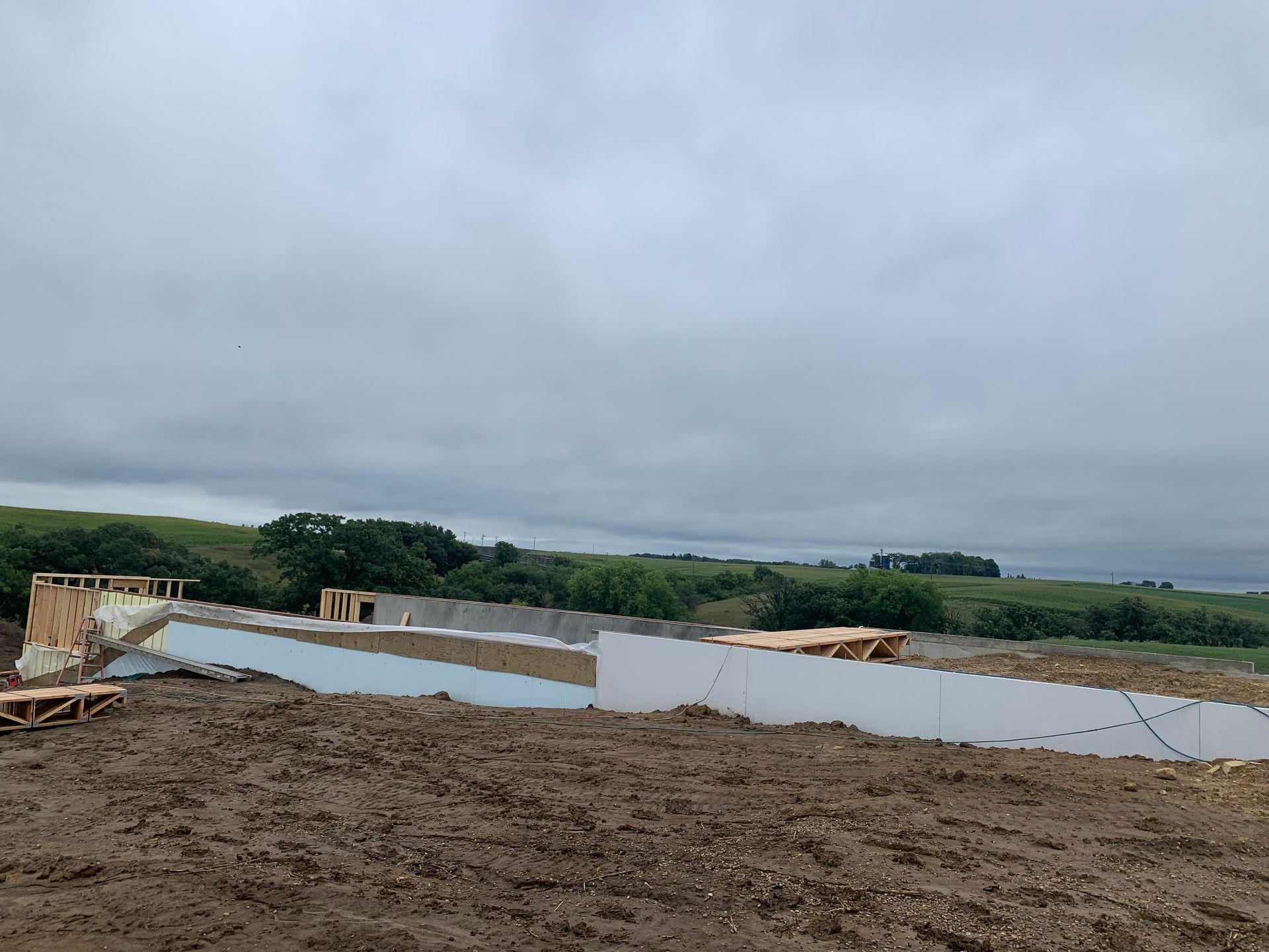 Foundation construction on a muddy site under a cloudy sky, with distant greenery.