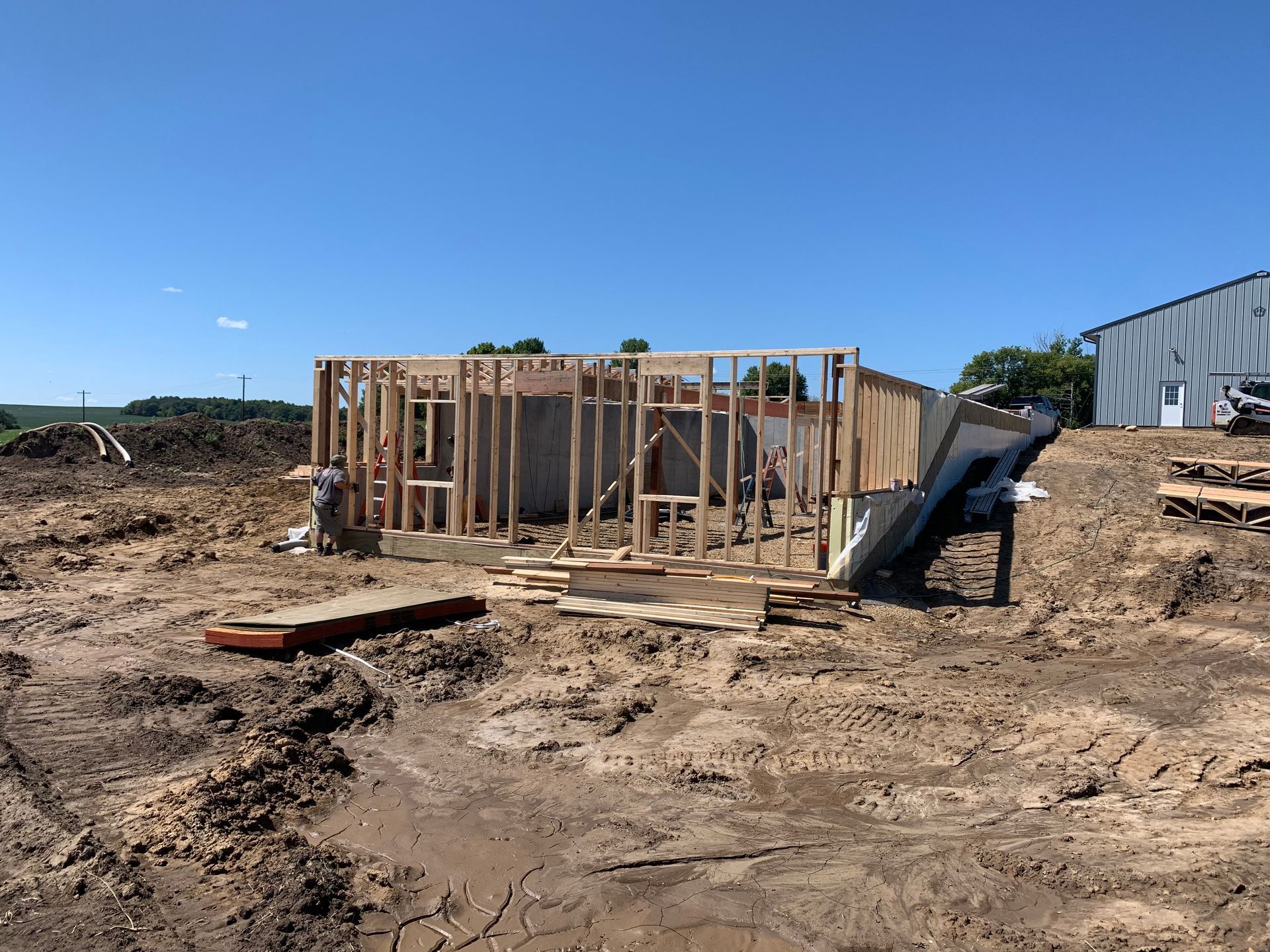 Wooden structure under construction on a dirt lot, with a building in the background under a blue sky.