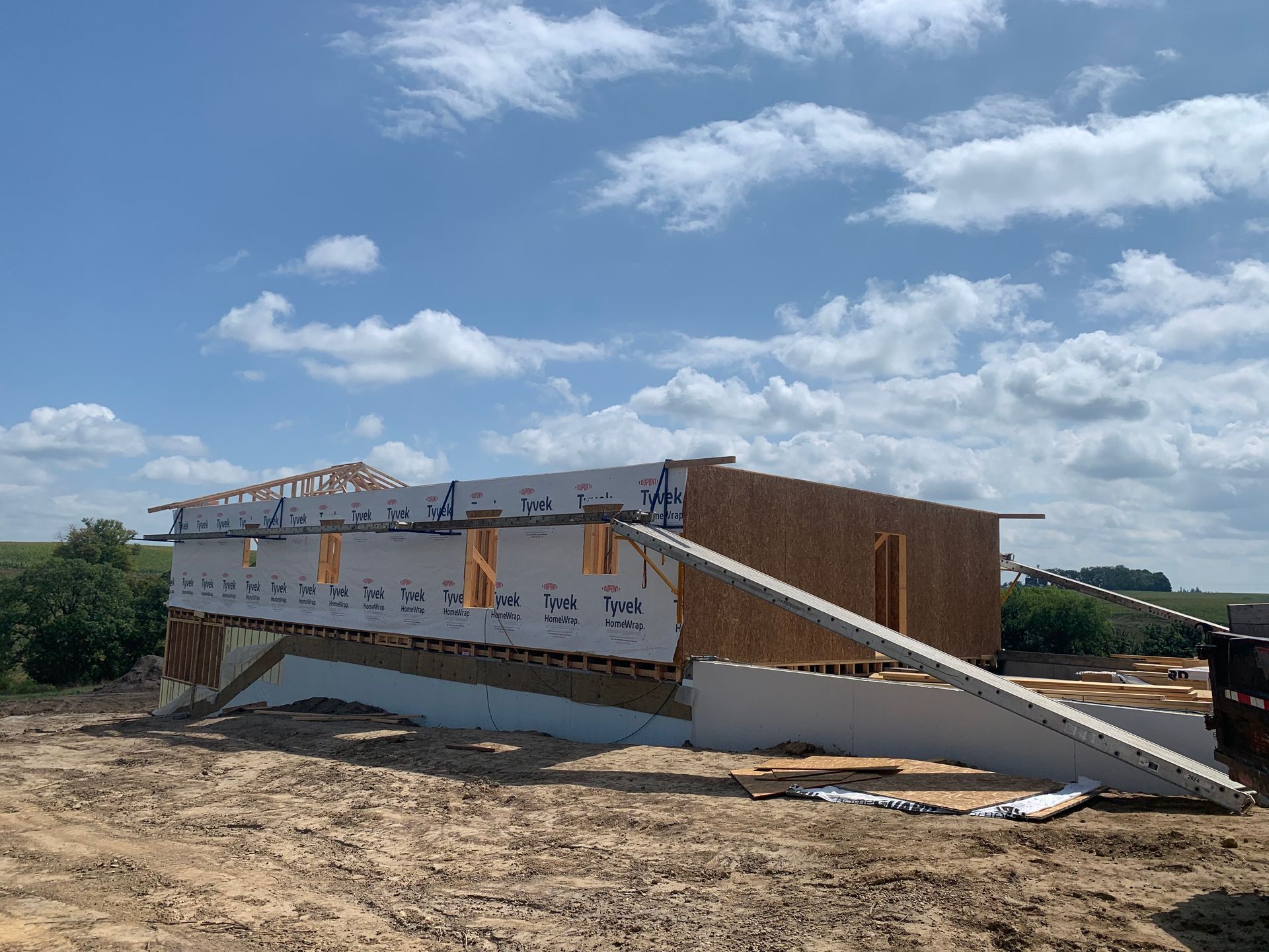 Construction site with a partially built house against a cloudy sky; visible wood framing, sheathing, and white wrapping.