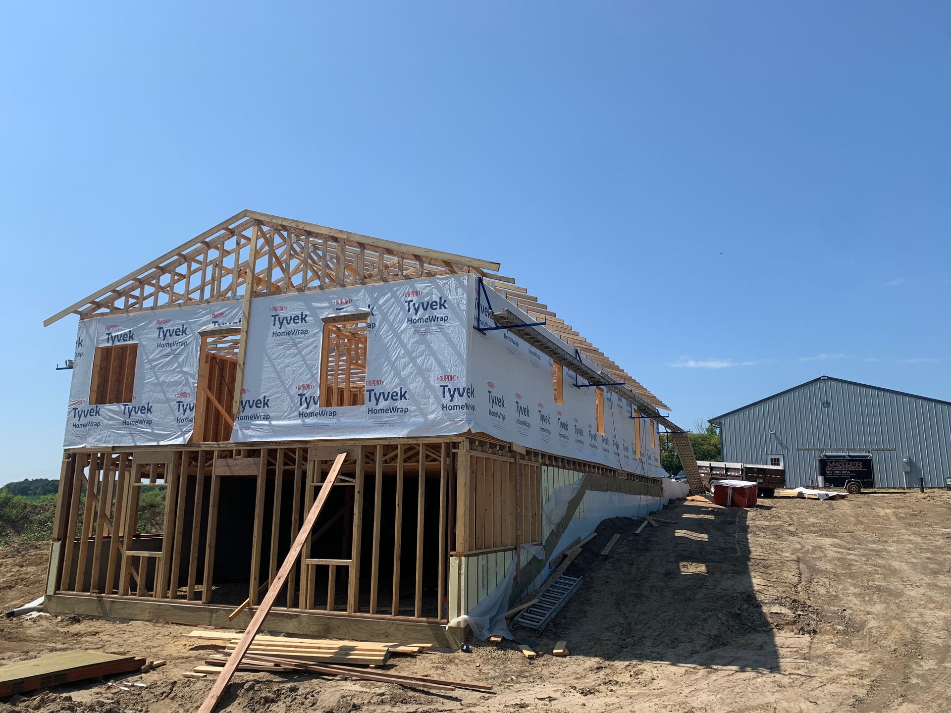 House under construction on a sunny day with wooden frame, blue sky, and a metal building in the distance.