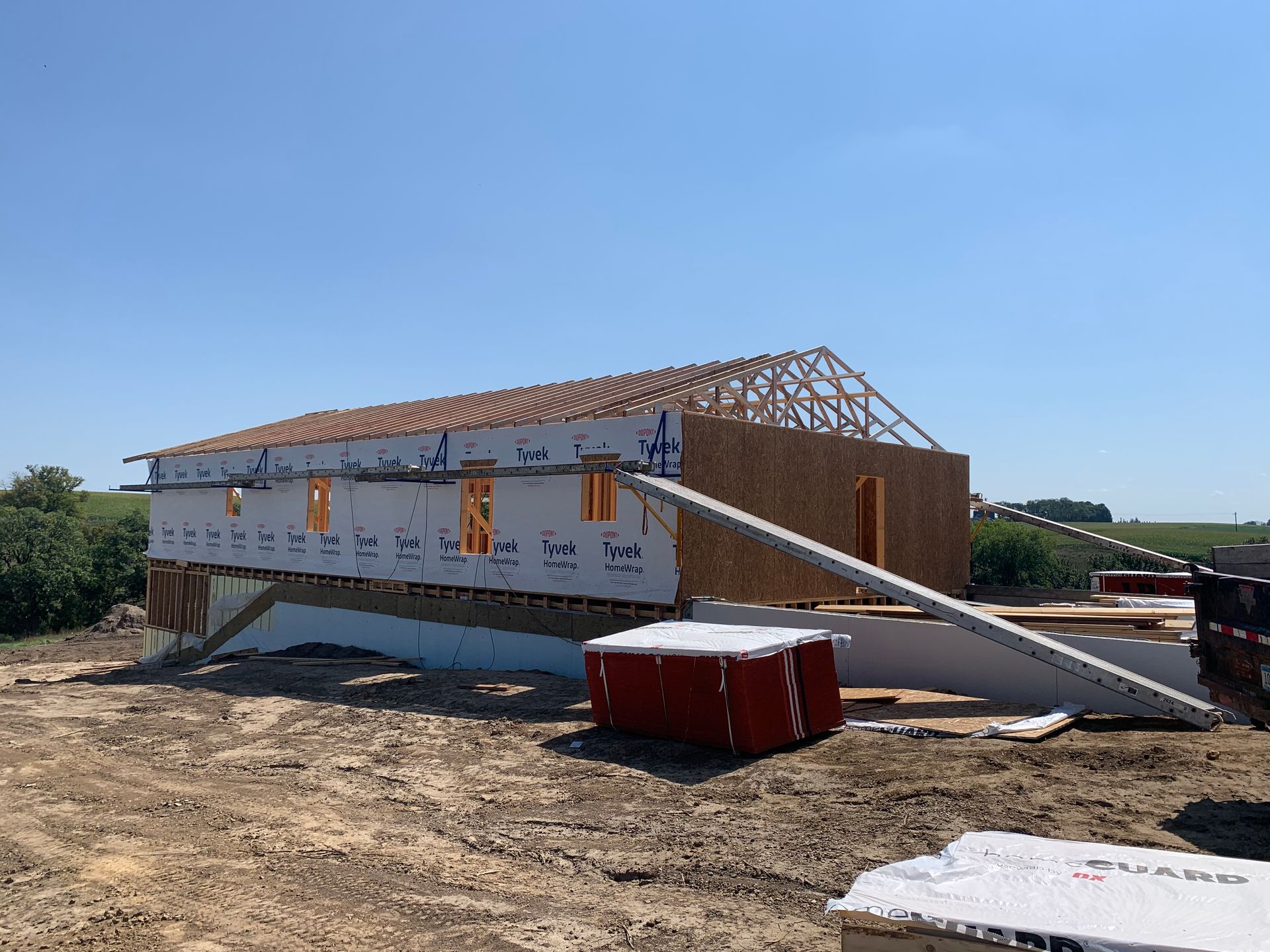 Construction site: A partially built house with exposed wood framing, blue wrap, and a gable roof under a clear blue sky.