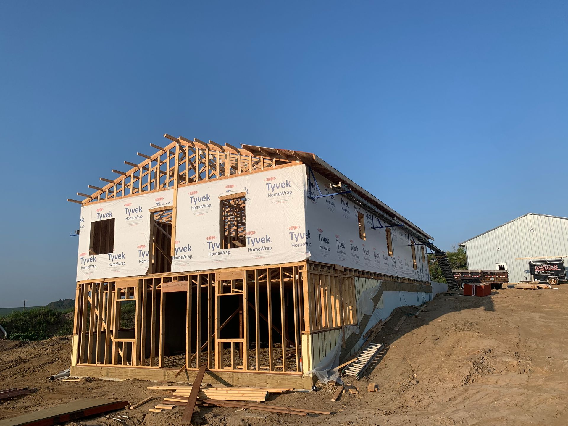 Two-story wooden house under construction on a hillside, with blue sky overhead.