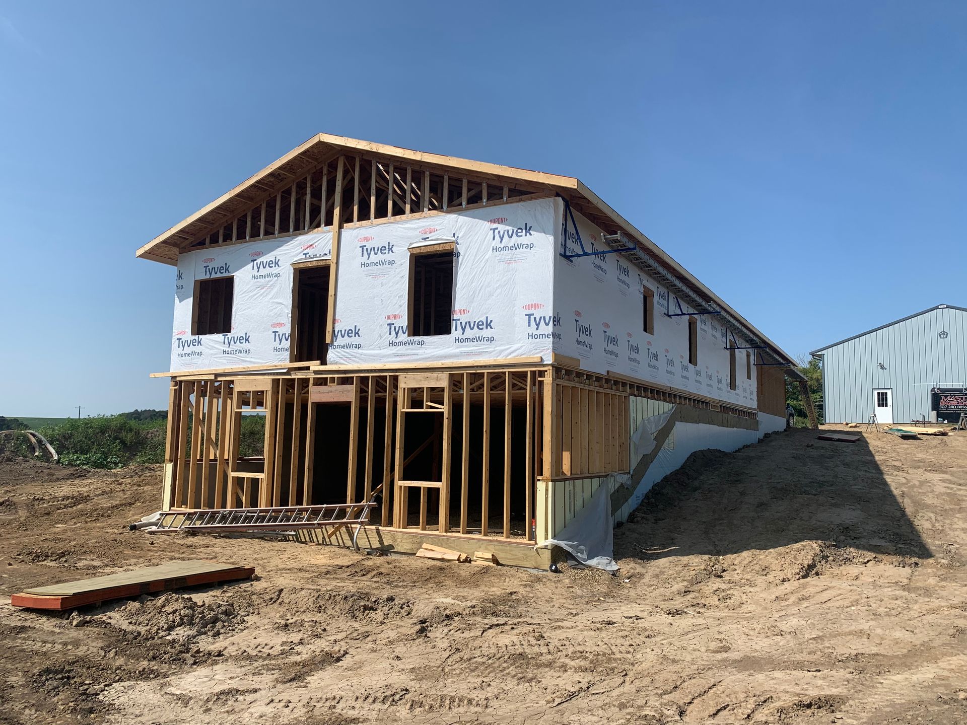 Two-story house under construction, wooden frame, wrapped in white material, set on a graded lot under a blue sky.