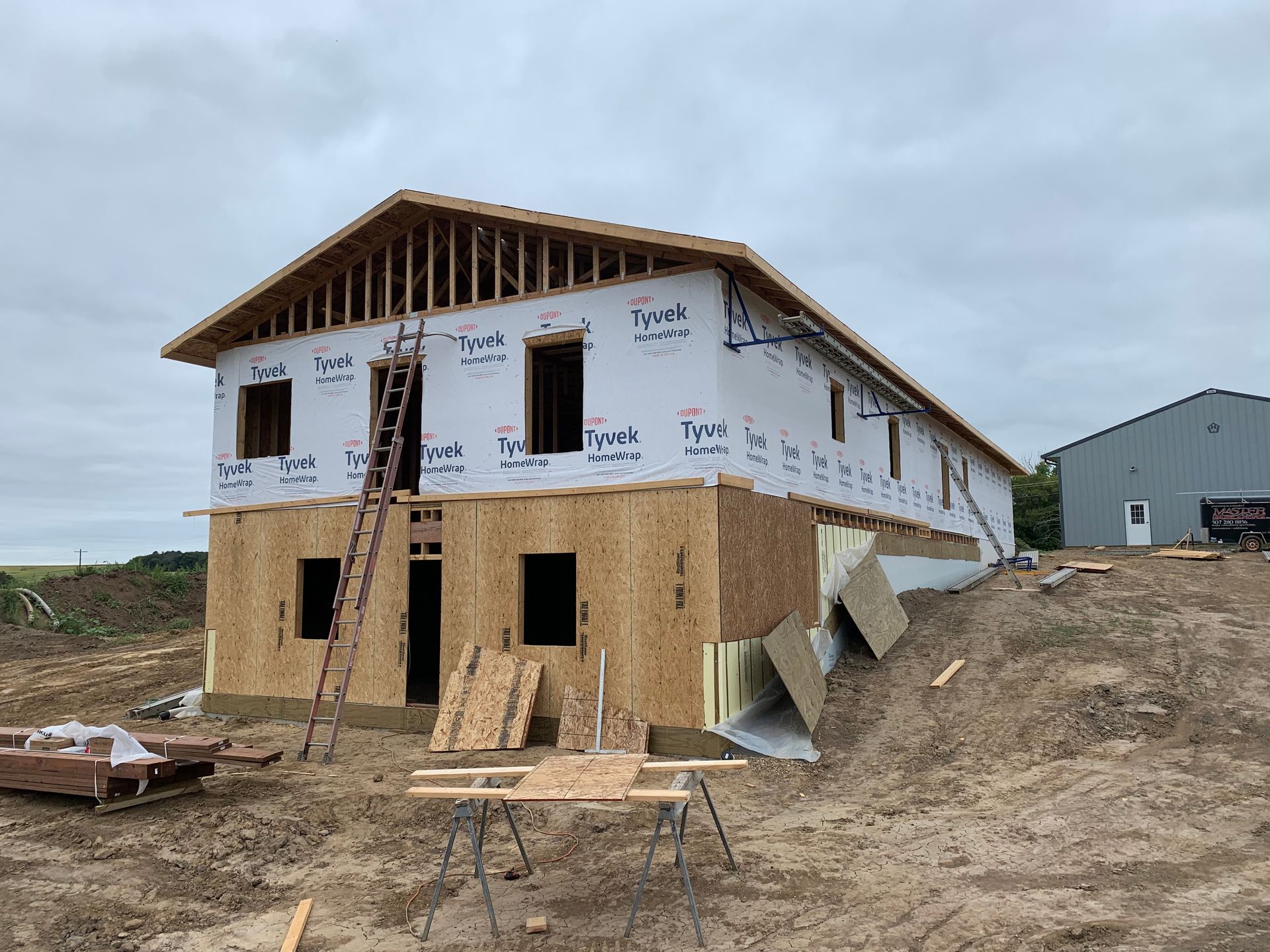 Two-story house under construction; wood framing with sheathing; insulation, ladder, and shed in the background.