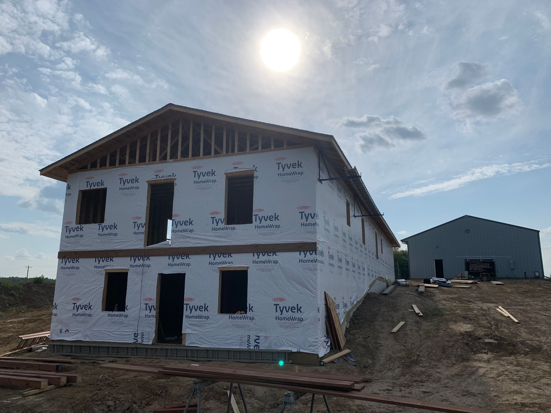 Two-story house under construction, wrapped in Tyvek, with a shed in the background, on a sunny day.