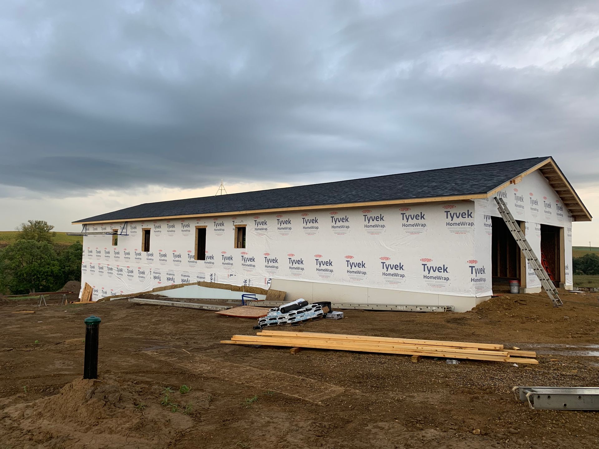A building under construction with white wrap, a dark roof, and windows, on a brown earth with a cloudy sky.
